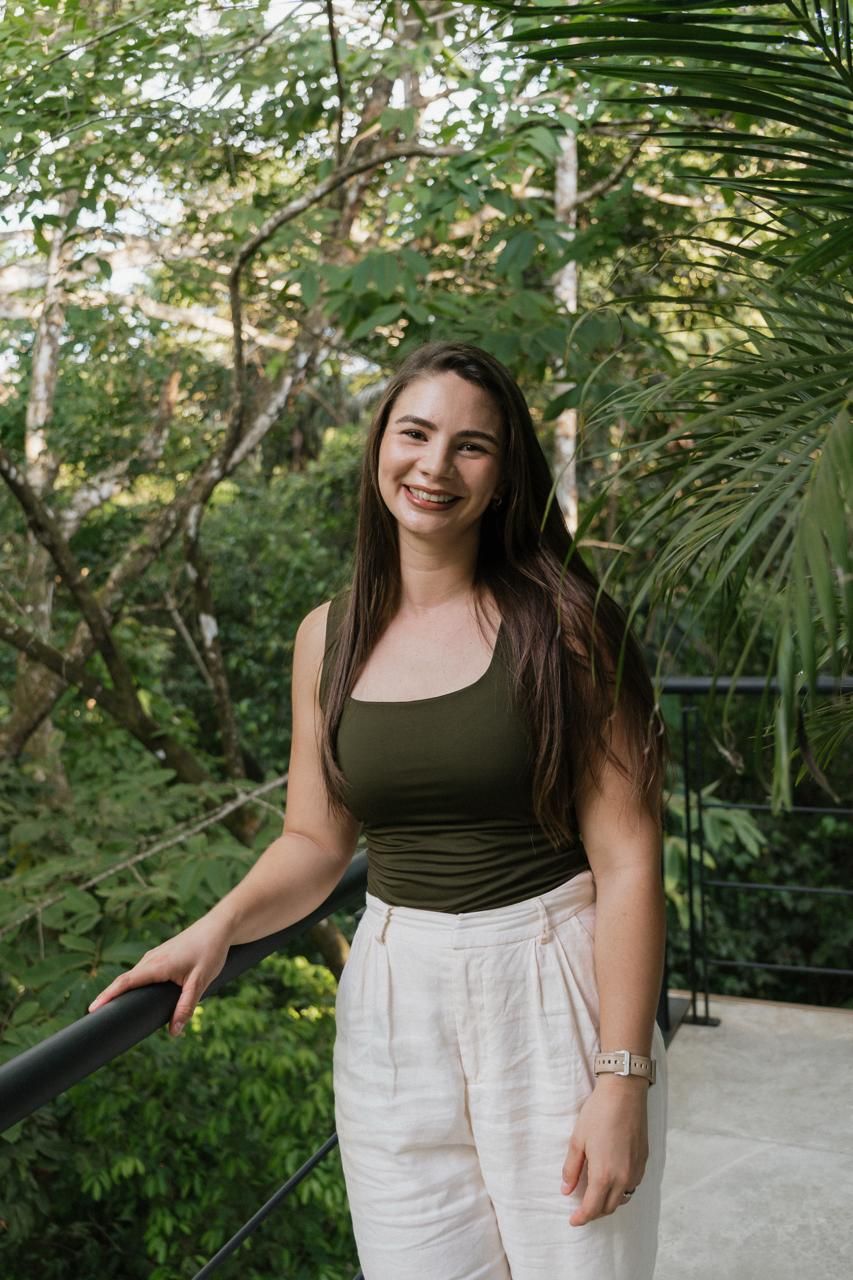 Smiling young woman in olive tank top and white linen pants standing on a balcony surrounded by lush greenery.