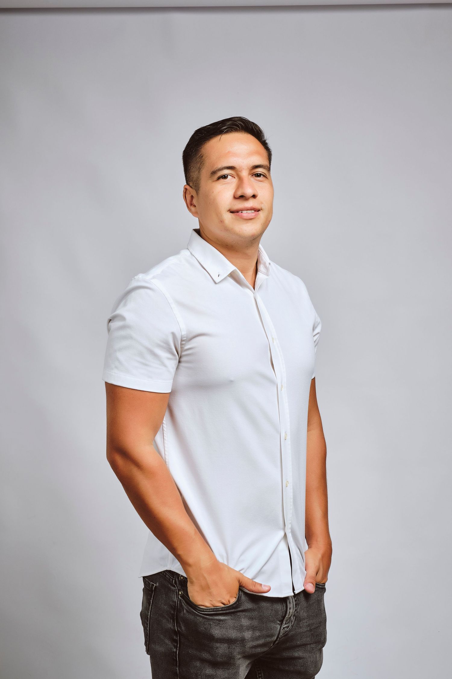 Young man in white short-sleeve button-up shirt and dark jeans posing confidently against a gray background.