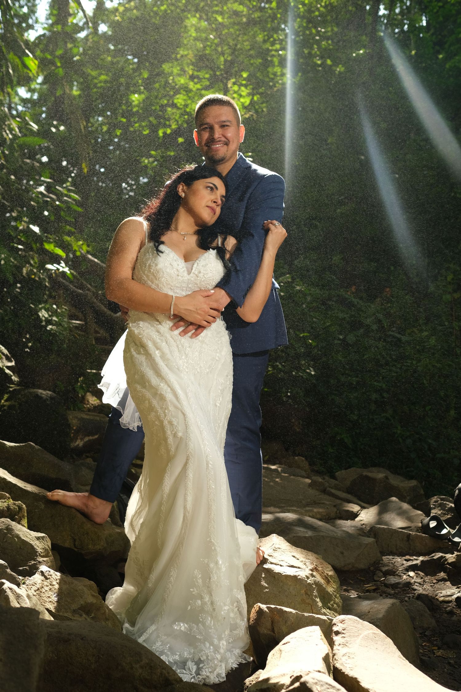 Wedding couple embraces on rocky terrain with sunbeams filtering through tropical forest greenery.