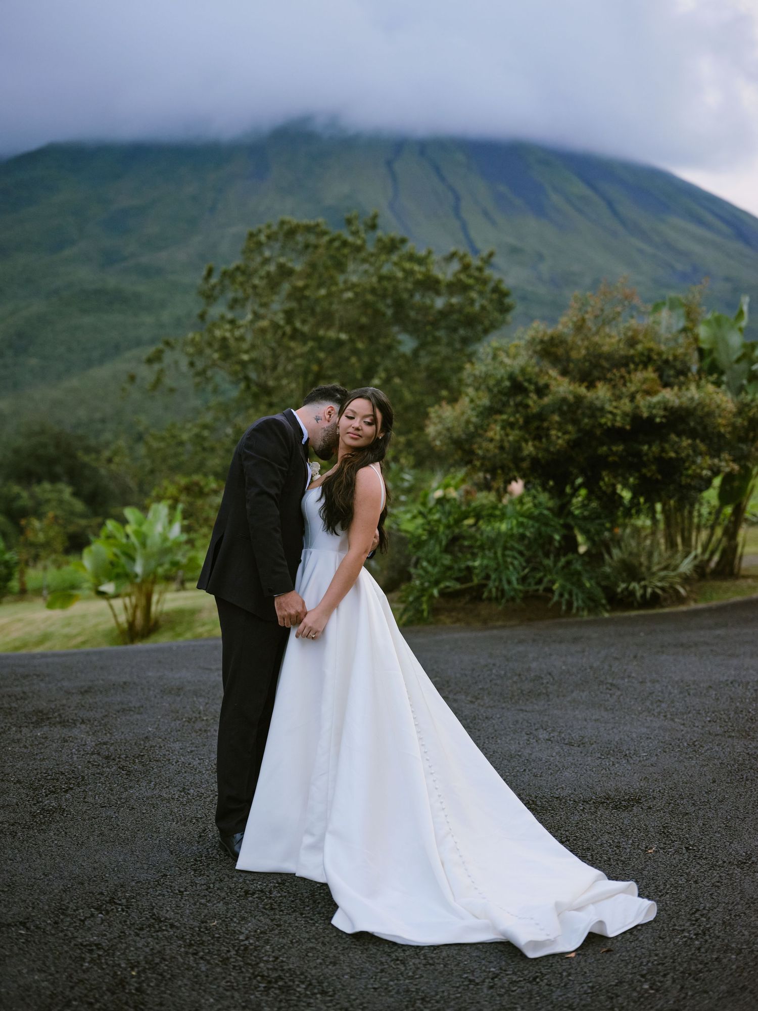 Couple posing romantically with dramatic volcanic mountain and cloudy sky in background.