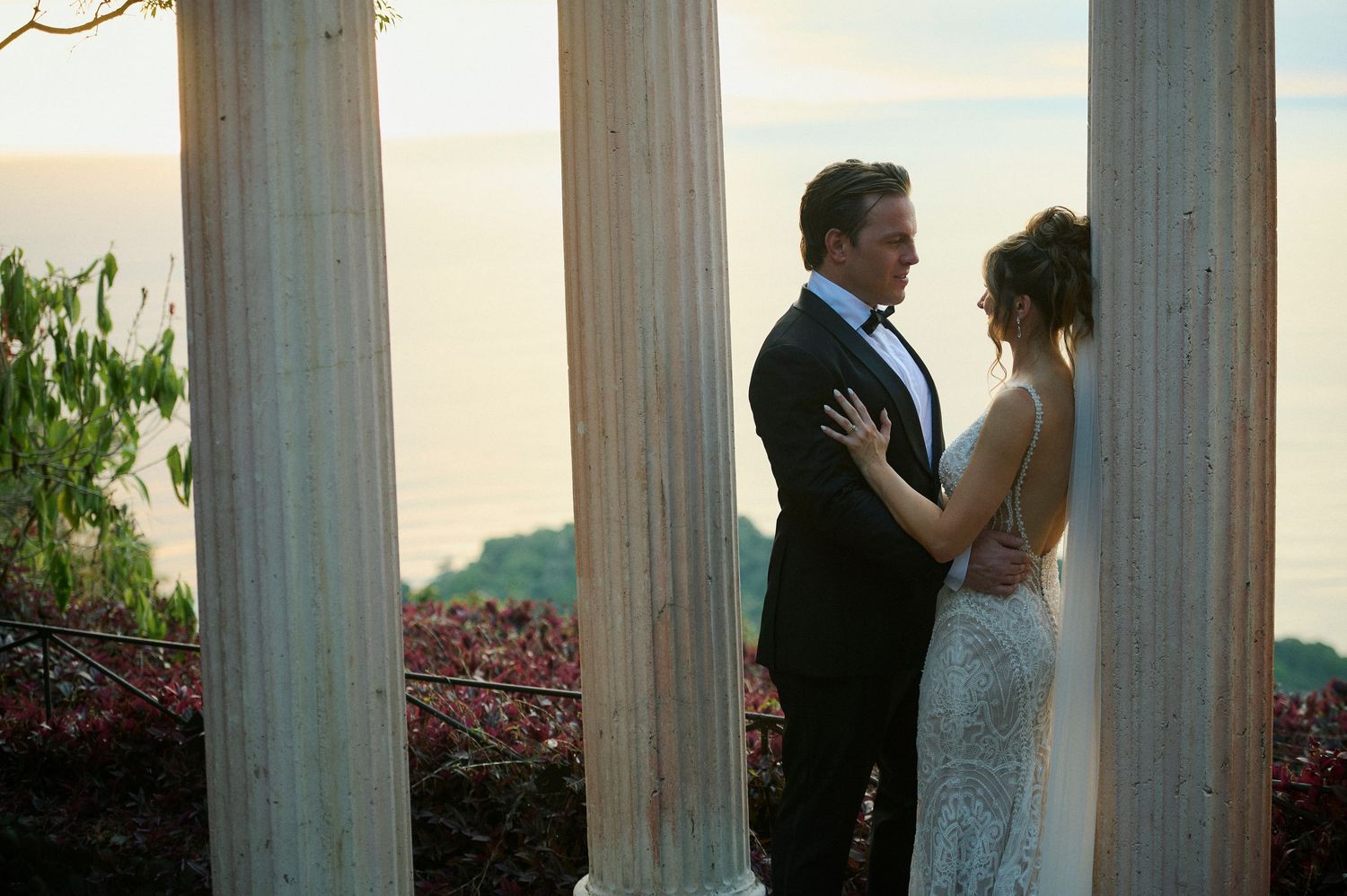 Romantic couple embracing between elegant white columns with ocean view at golden hour wedding.
