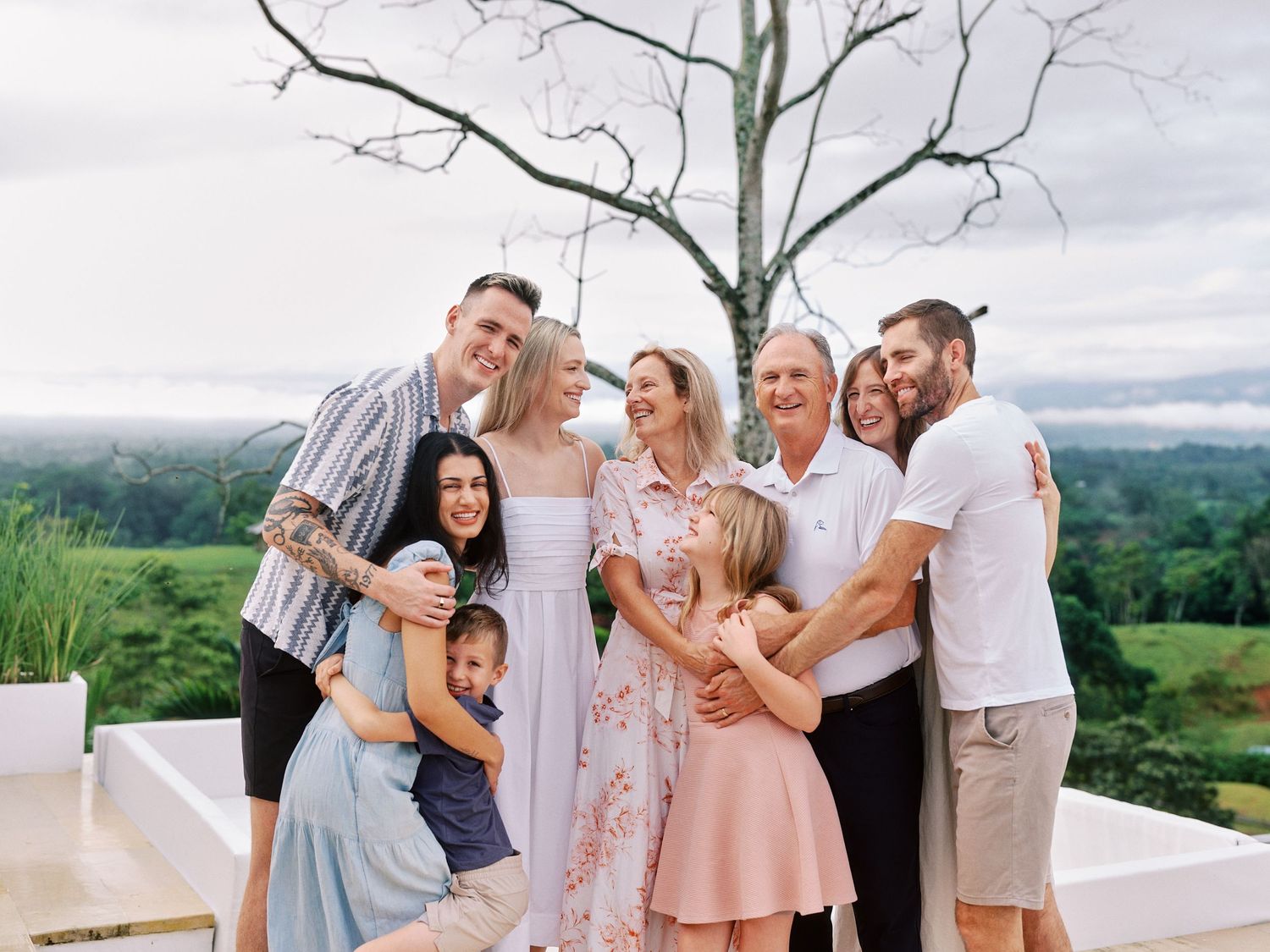 Extended family sharing warm embraces while posing together on an outdoor terrace with tropical landscape views.