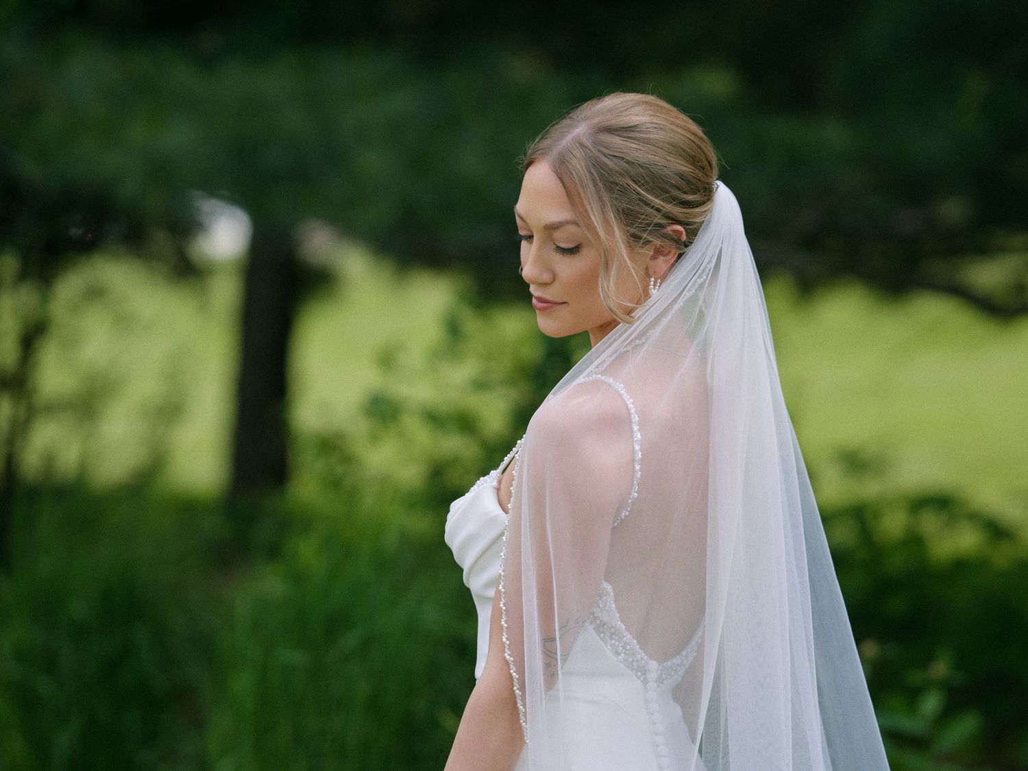 Beautiful bride wearing a flowing white veil and wedding gown standing in a lush green outdoor setting.