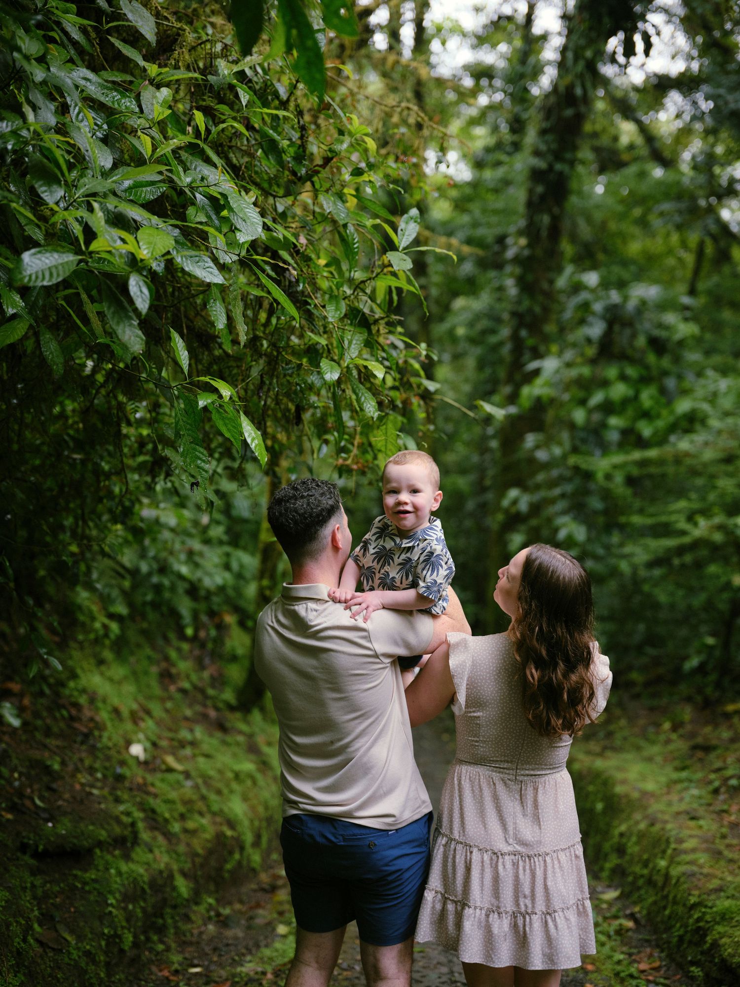 Family of three with baby exploring a lush tropical rainforest path surrounded by green vegetation.