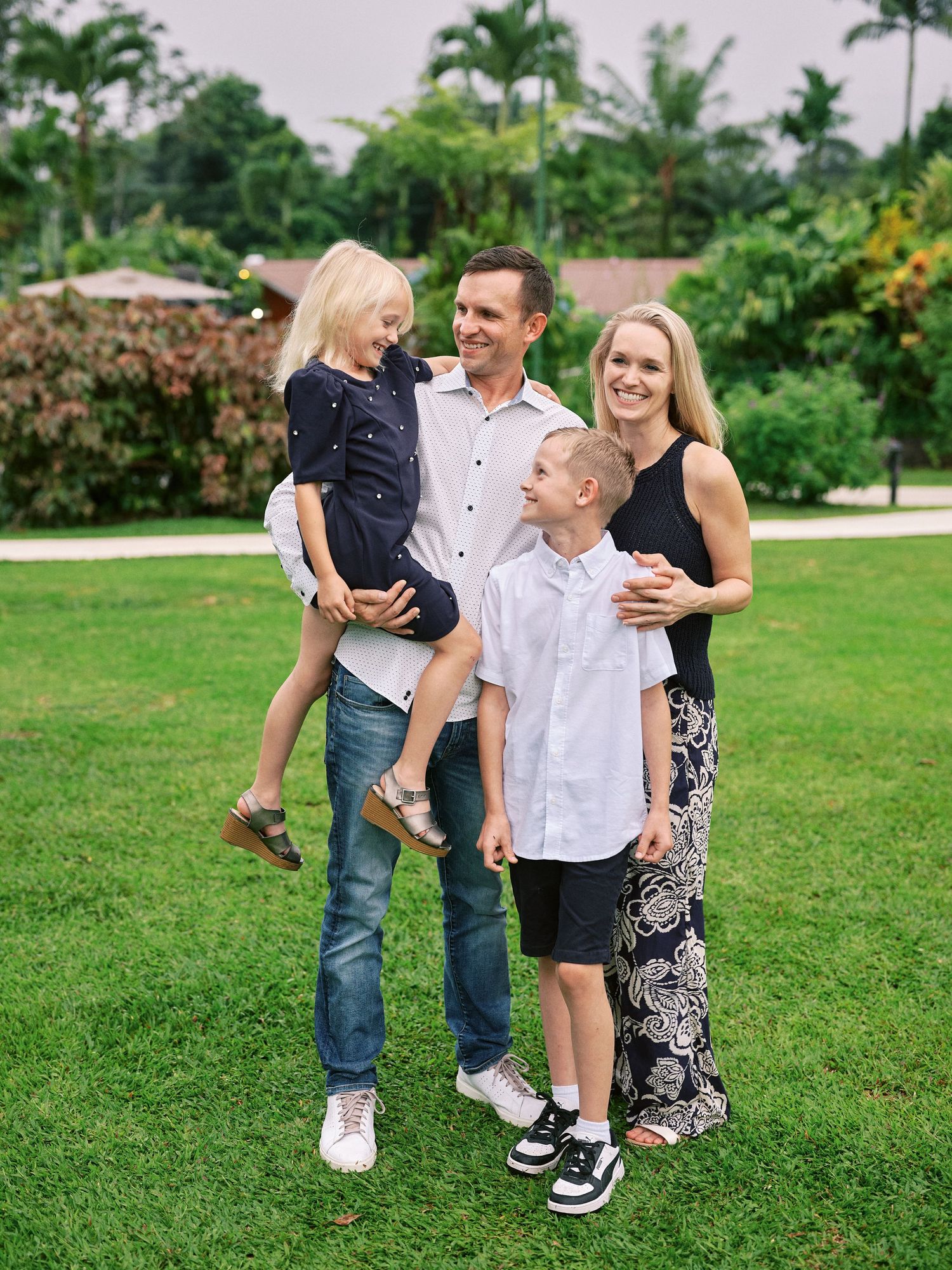 Happy family of four posing on green lawn with tropical trees, parents holding young children in coordinated navy outfits.