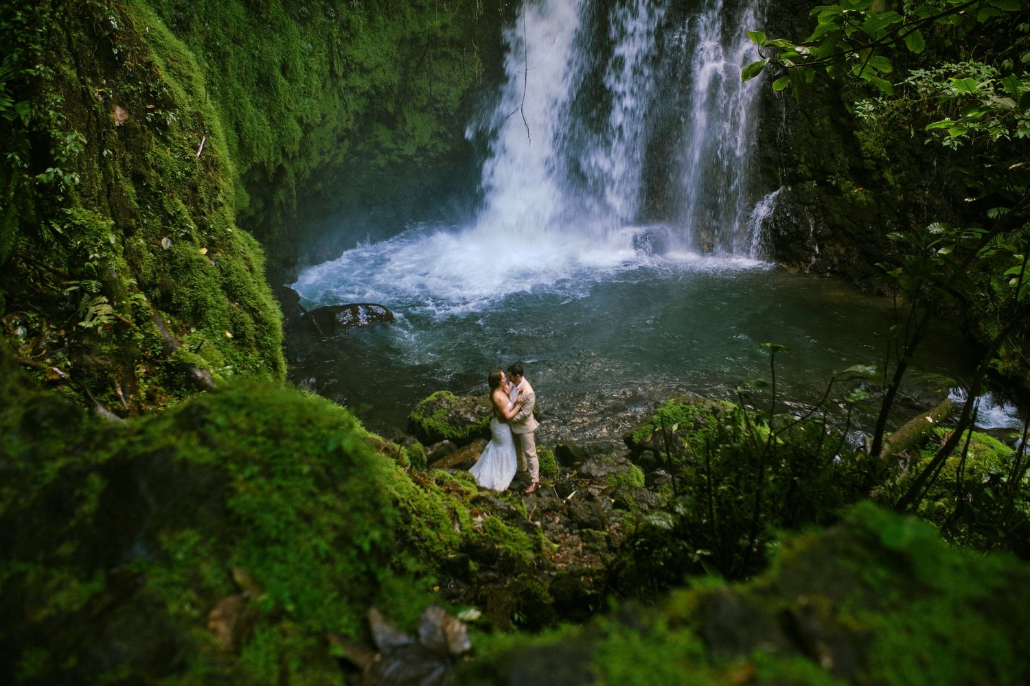 Couple embracing romantically at the base of a lush waterfall surrounded by green moss.