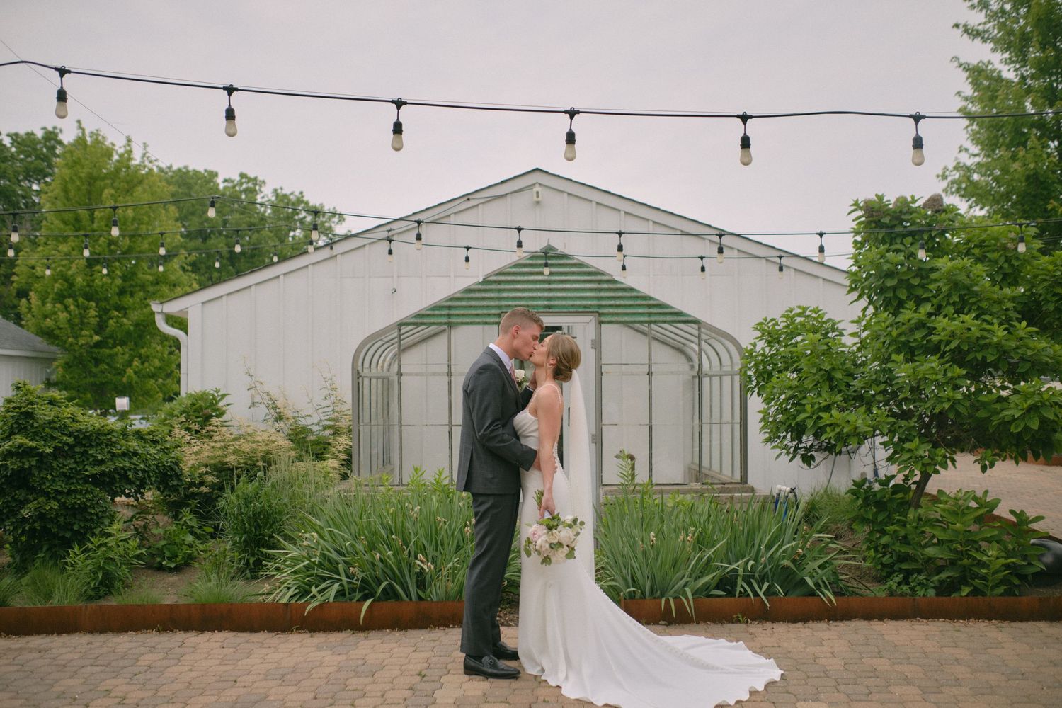 Bride and groom sharing a tender moment in front of a charming greenhouse with string lights overhead.