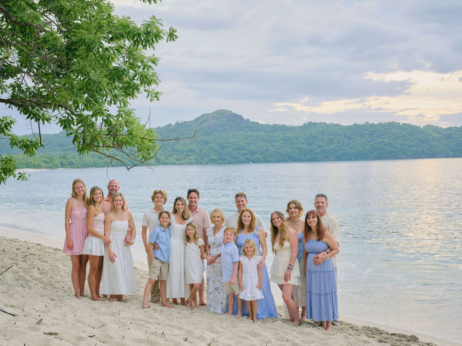 Large family group posing together on a tropical beach with lush green mountains and calm water in the background.