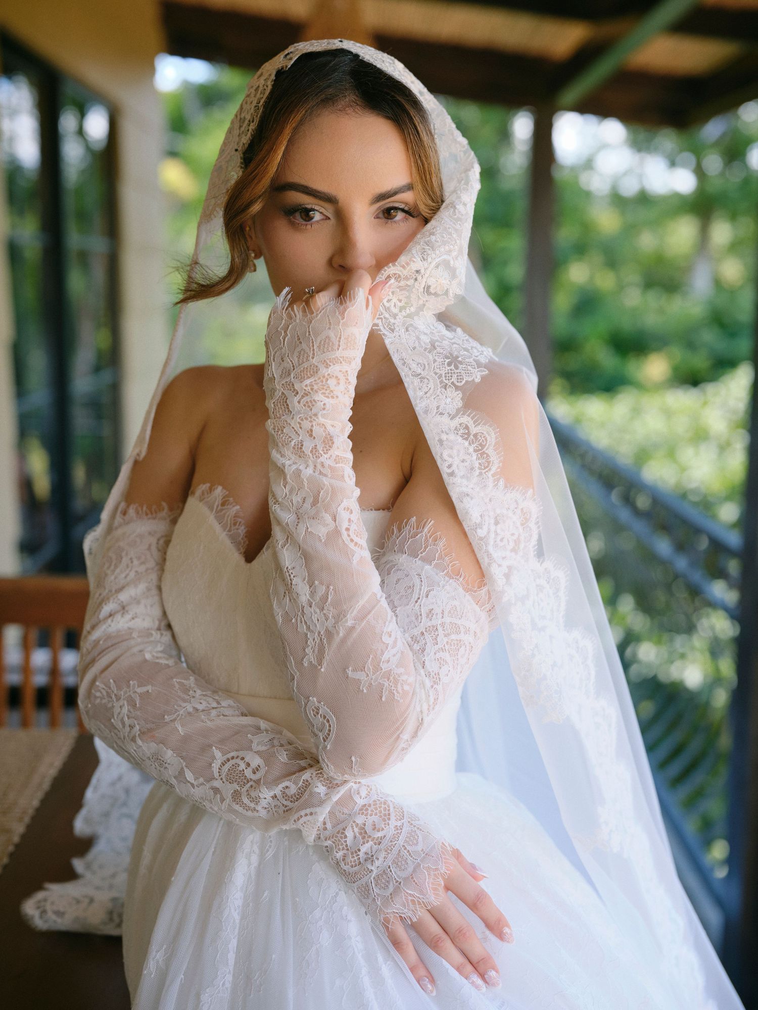 Bride in lace gloves and veil poses gracefully on a balcony surrounded by tropical foliage.