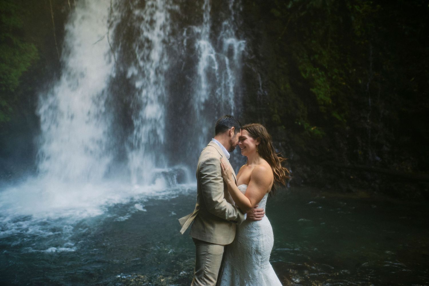Bride and groom sharing a romantic kiss in front of a stunning misty waterfall.