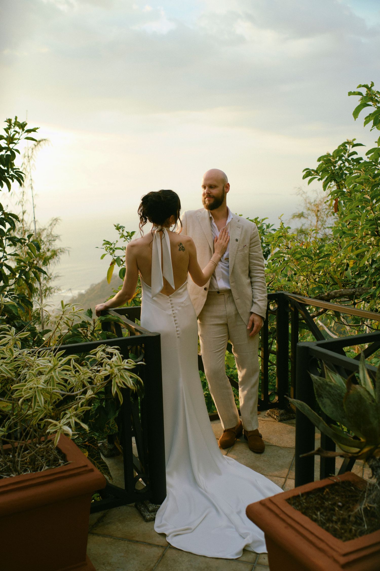 Bride in backless gown and groom enjoy a scenic balcony view surrounded by tropical greenery.