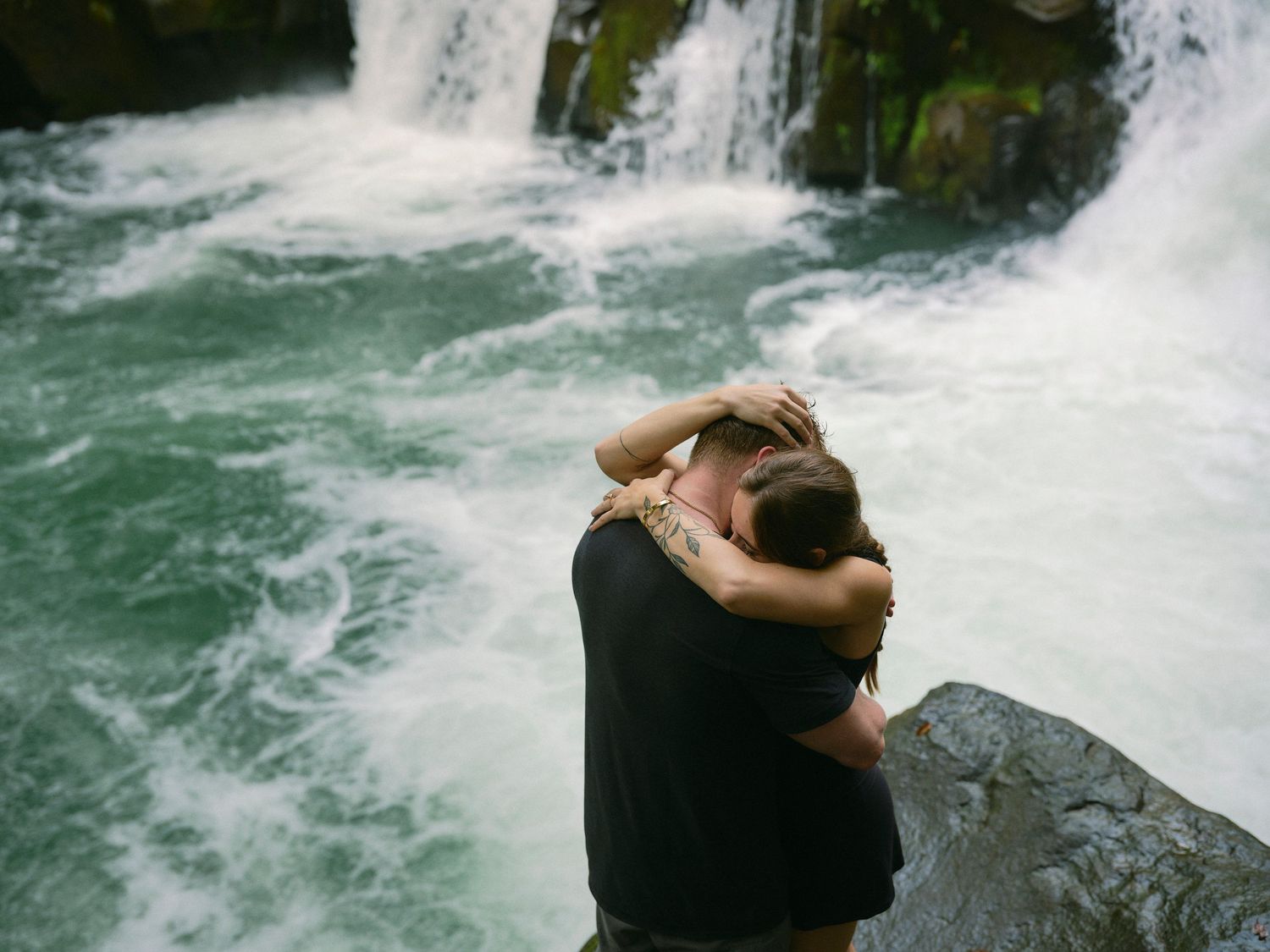 Couple embracing romantically on rocks in front of a rushing waterfall in a lush outdoor setting.