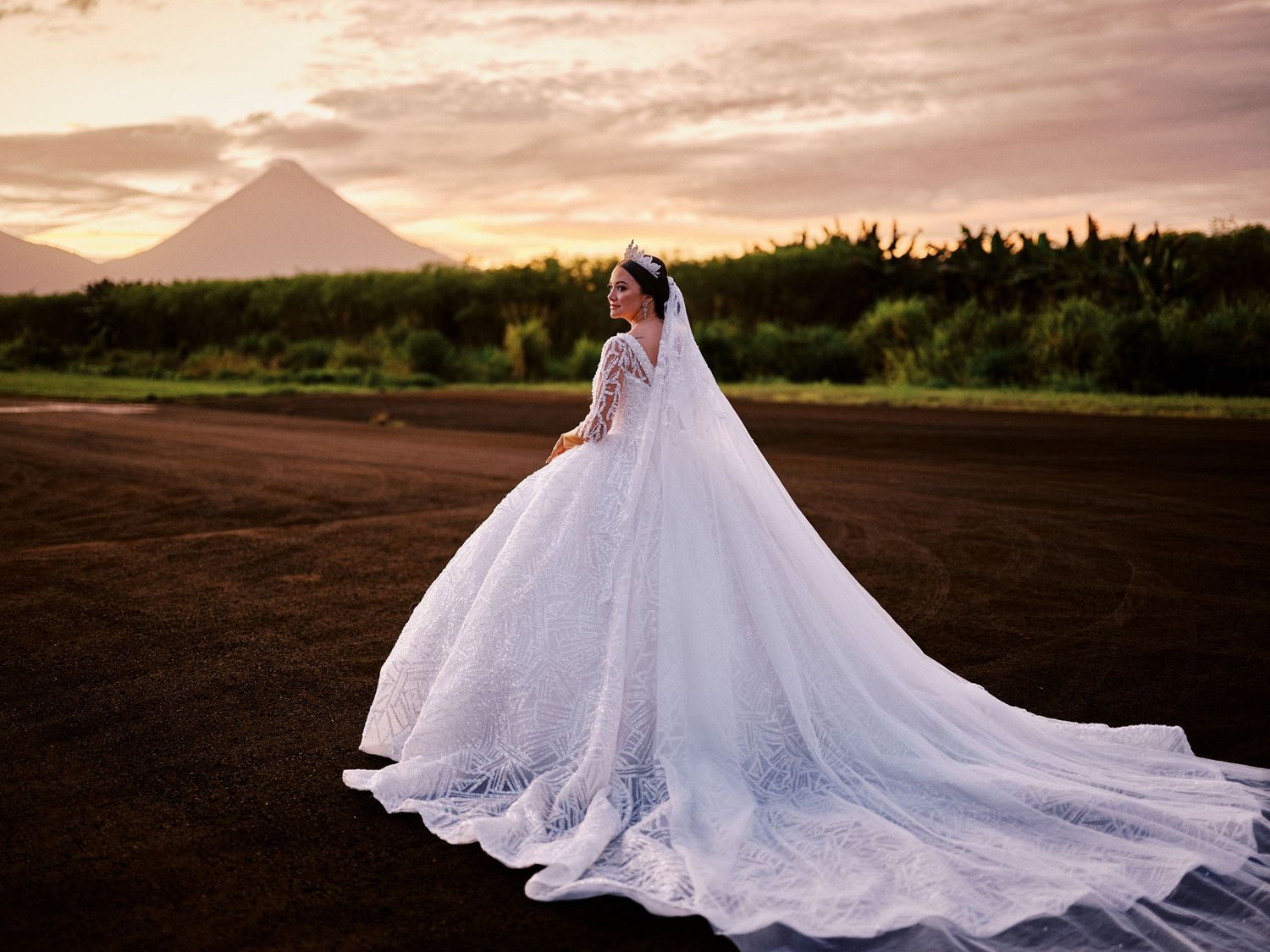 Bride in a stunning ball gown with long train poses before a volcanic mountain at sunset.