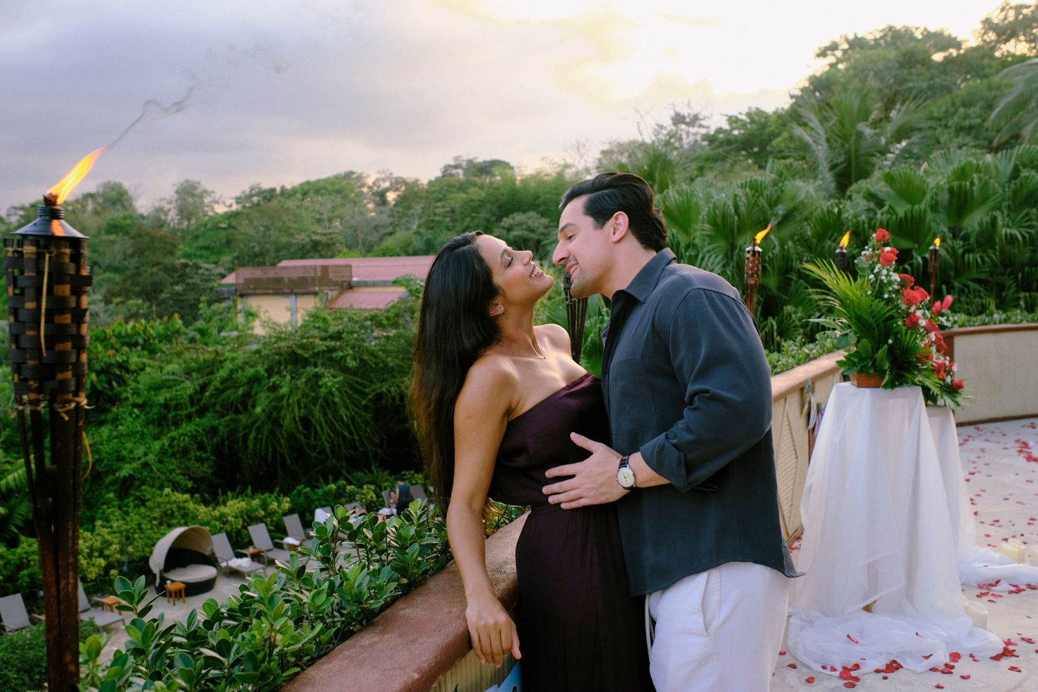 Newly engaged couple embracing tenderly on a tropical terrace at dusk with lush greenery in background.