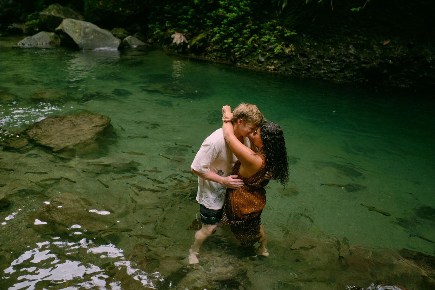 Couple embracing and kissing while standing in a serene green river surrounded by lush forest rocks.