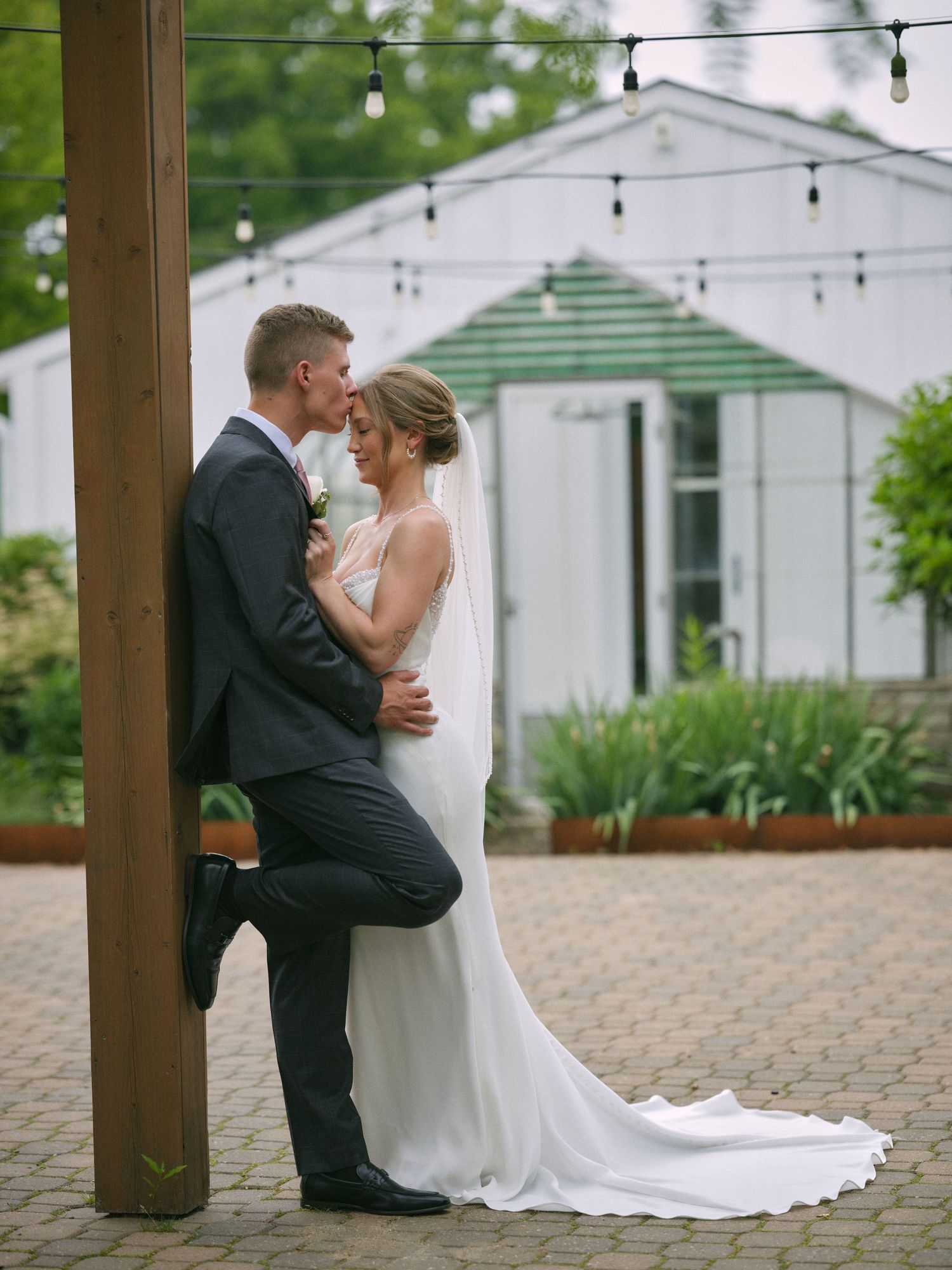 Bride and groom sharing a kiss while leaning against a wooden post with a greenhouse backdrop.