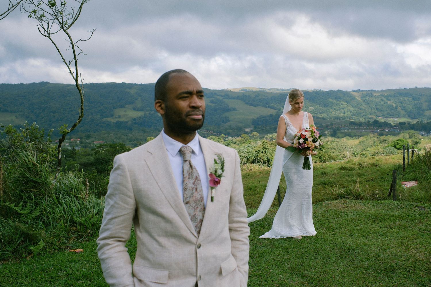 Groom in beige suit standing before bride in white gown at a lush green hillside wedding.