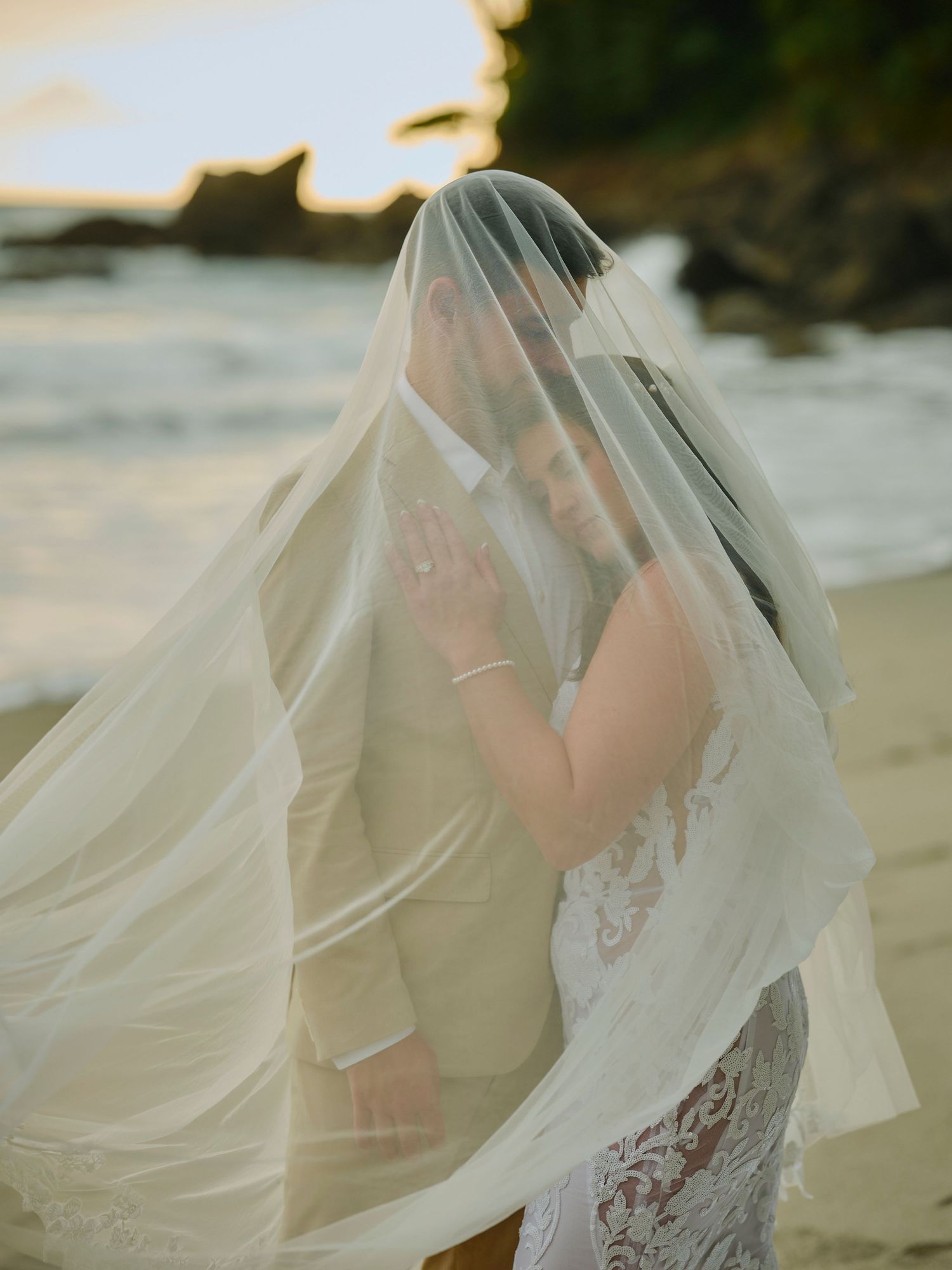 Bride and groom sharing kiss beneath flowing veil on sandy beach at sunset.