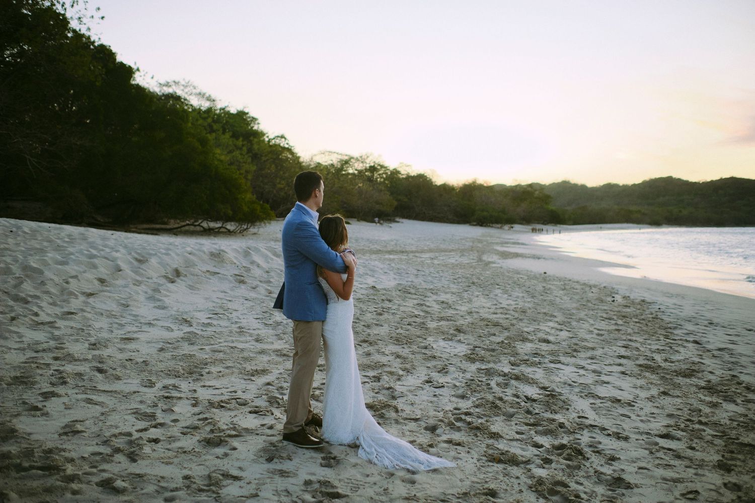 Wedding couple embracing on a white sandy beach at sunset with lush trees in background.