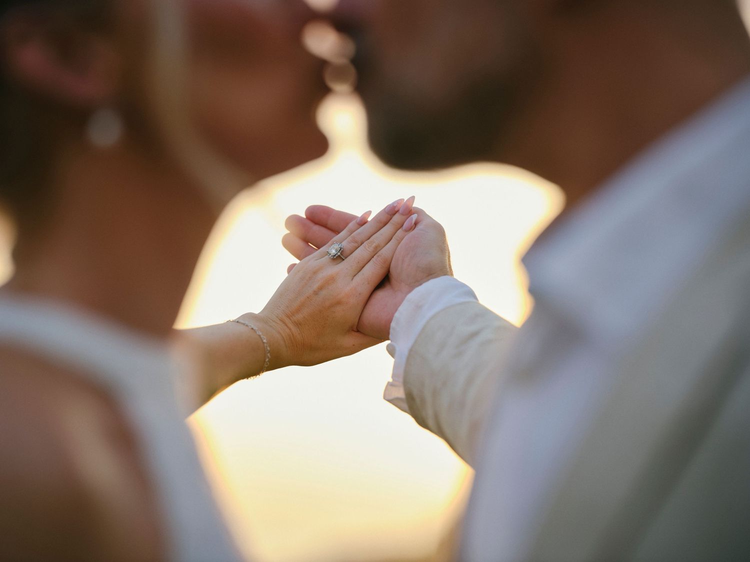Close-up of bride and groom holding hands showing wedding ring with warm golden sunset light glowing behind them.