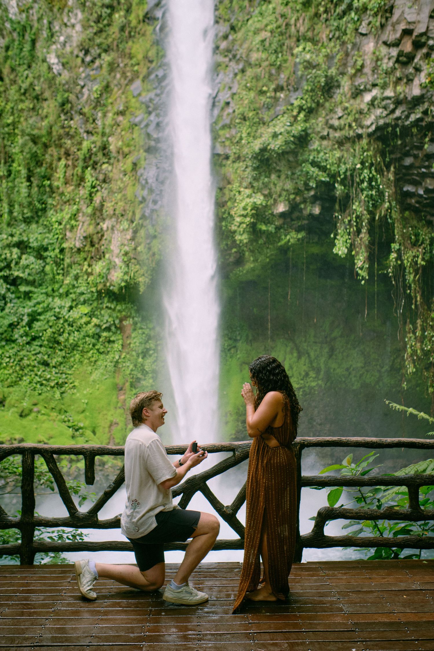 Man proposing on one knee to woman on wooden deck in front of tropical waterfall surrounded by greenery.