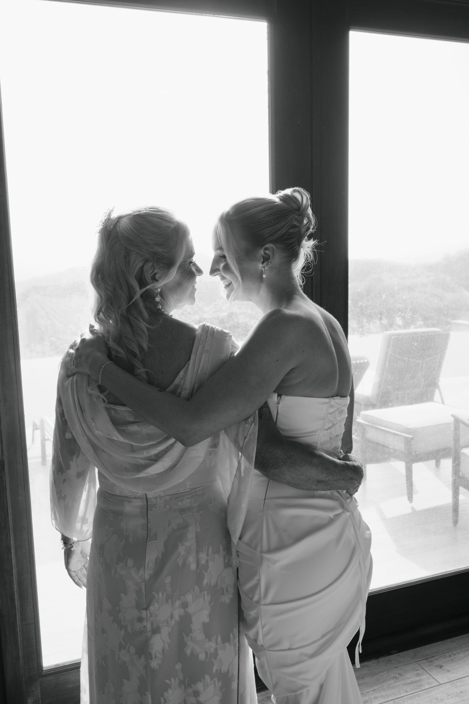 Two women in elegant dresses embrace and smile at each other in a black and white wedding photo.