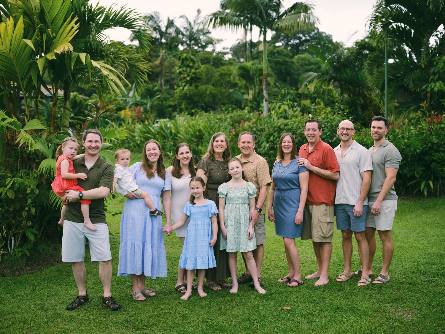 Large extended family group smiling together outdoors surrounded by vibrant tropical plants and palm trees.