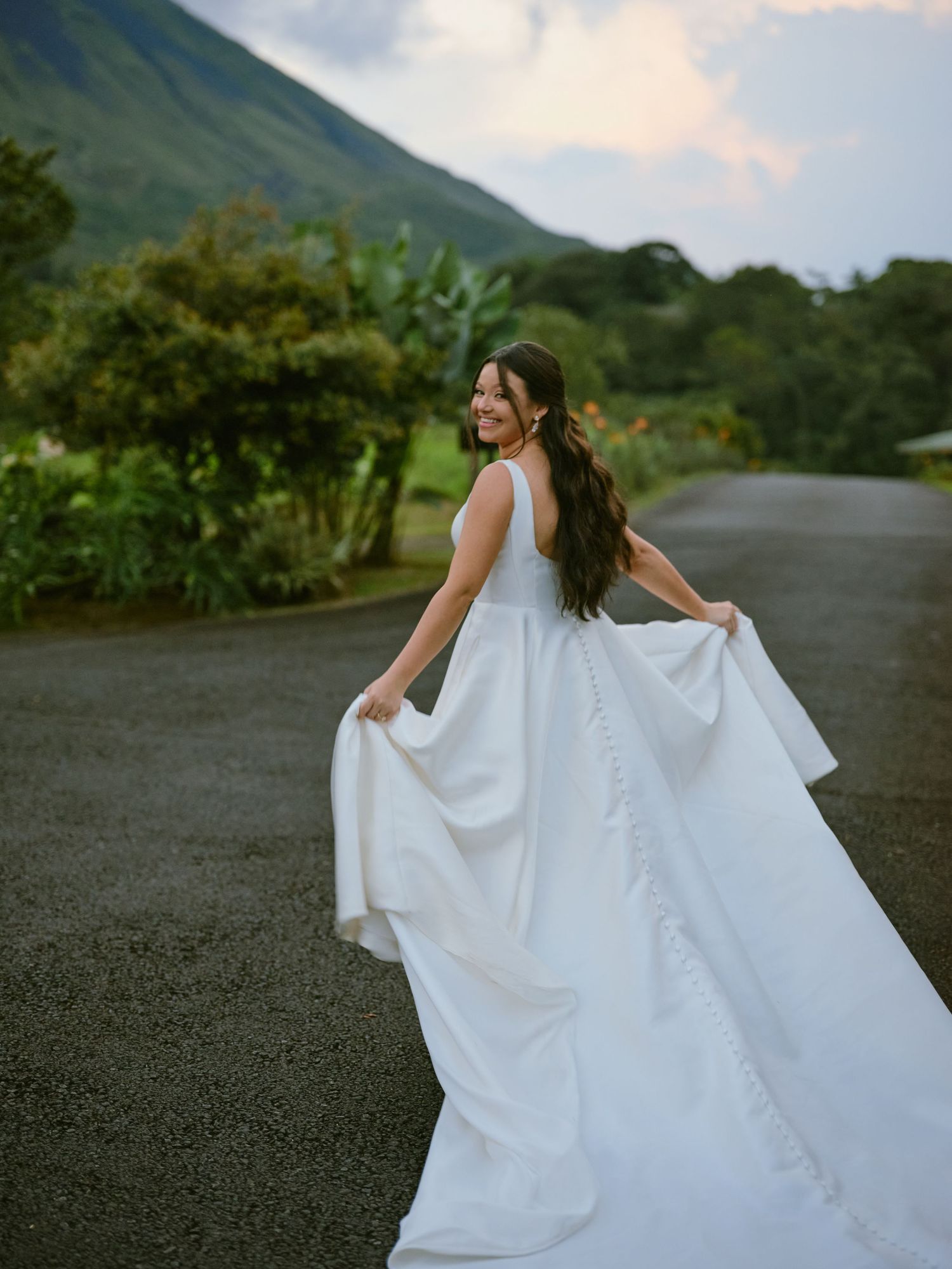 Bride twirling elegant white gown on road with lush volcanic mountain landscape behind.