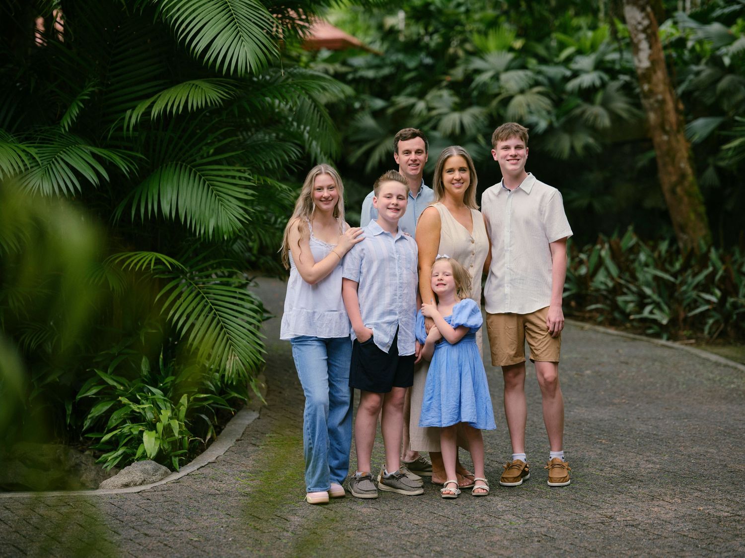Family of six smiling together on a lush tropical garden path surrounded by palm trees and green foliage.