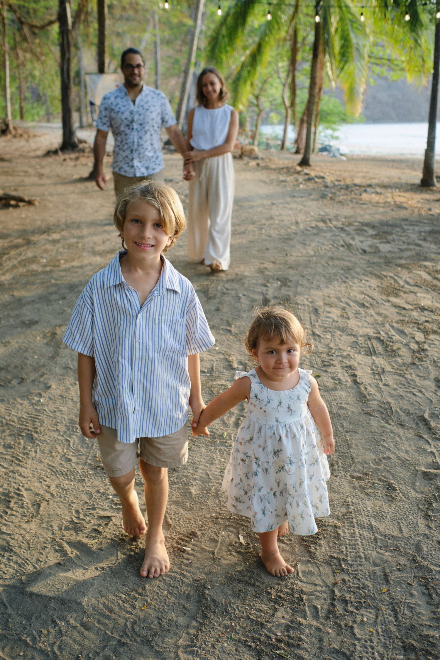 Two young children holding hands on sandy beach with parents standing under palm trees in background.