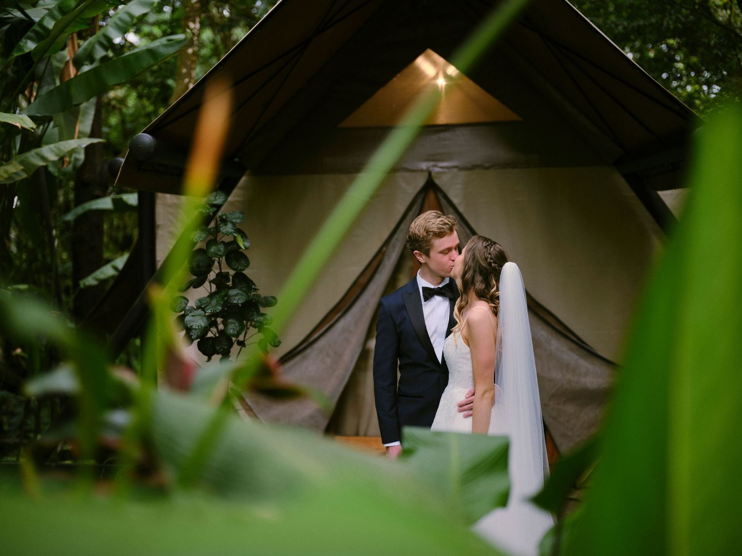 Newlyweds share a romantic kiss in front of a glamping tent nestled among tropical greenery.