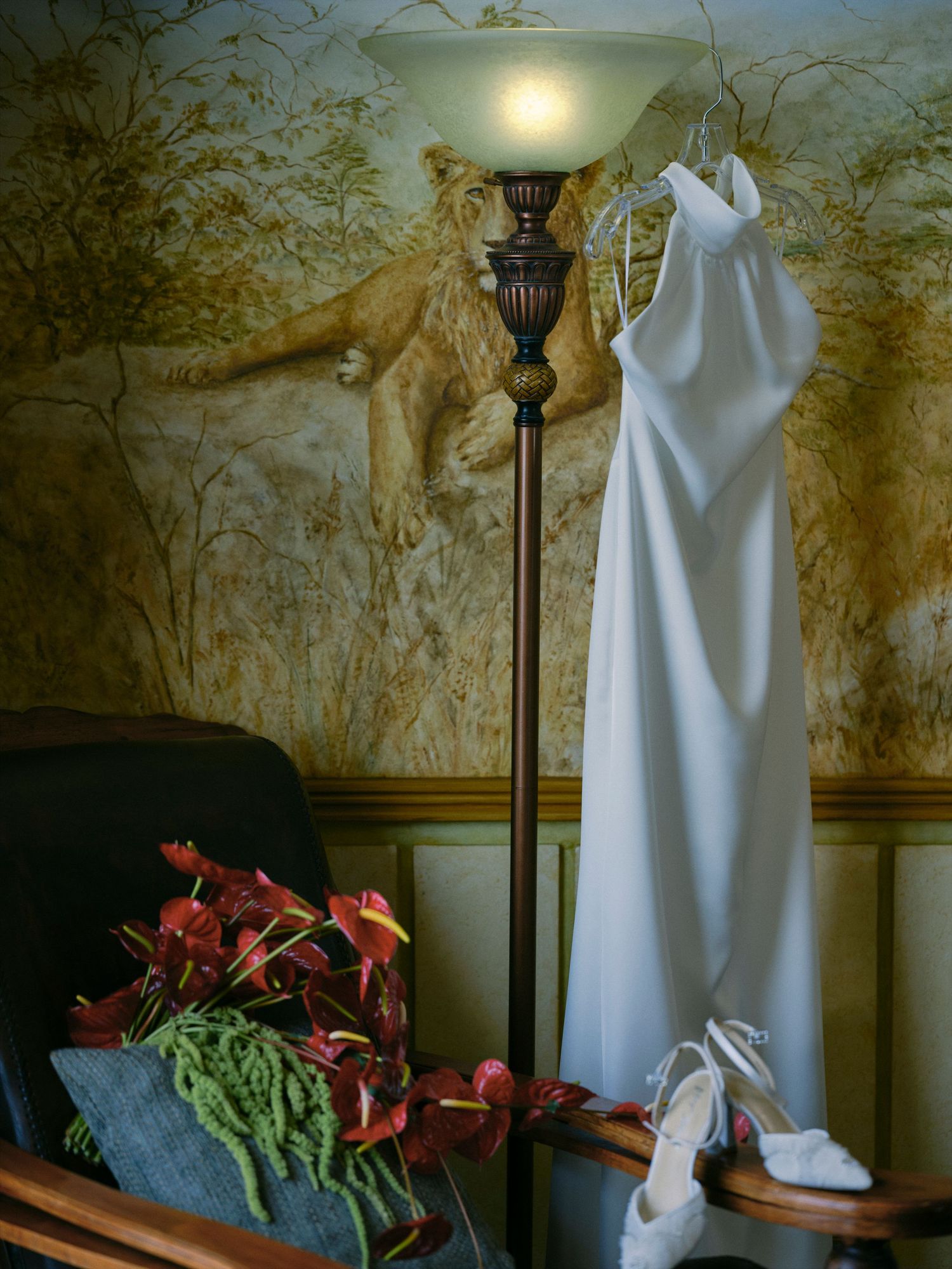 White halter wedding gown hangs on a lamp beside red flowers and heels in a safari-themed room.