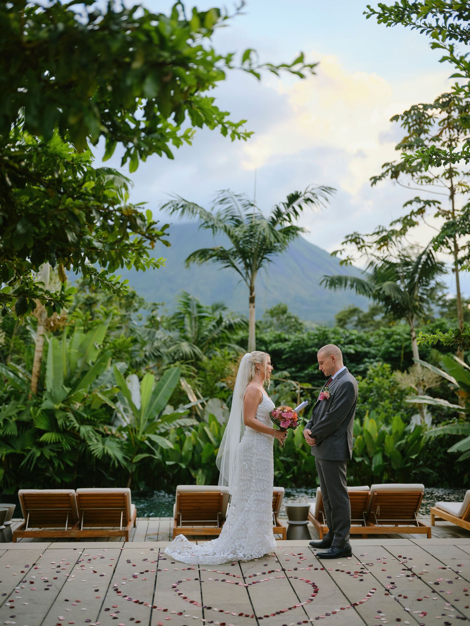 Bride and groom holding hands during outdoor ceremony with lush tropical garden and volcano in background.