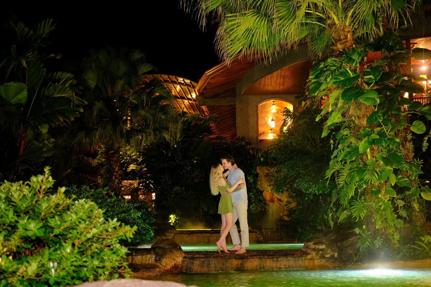 Couple shares romantic embrace surrounded by tropical palms and garden lights at night.