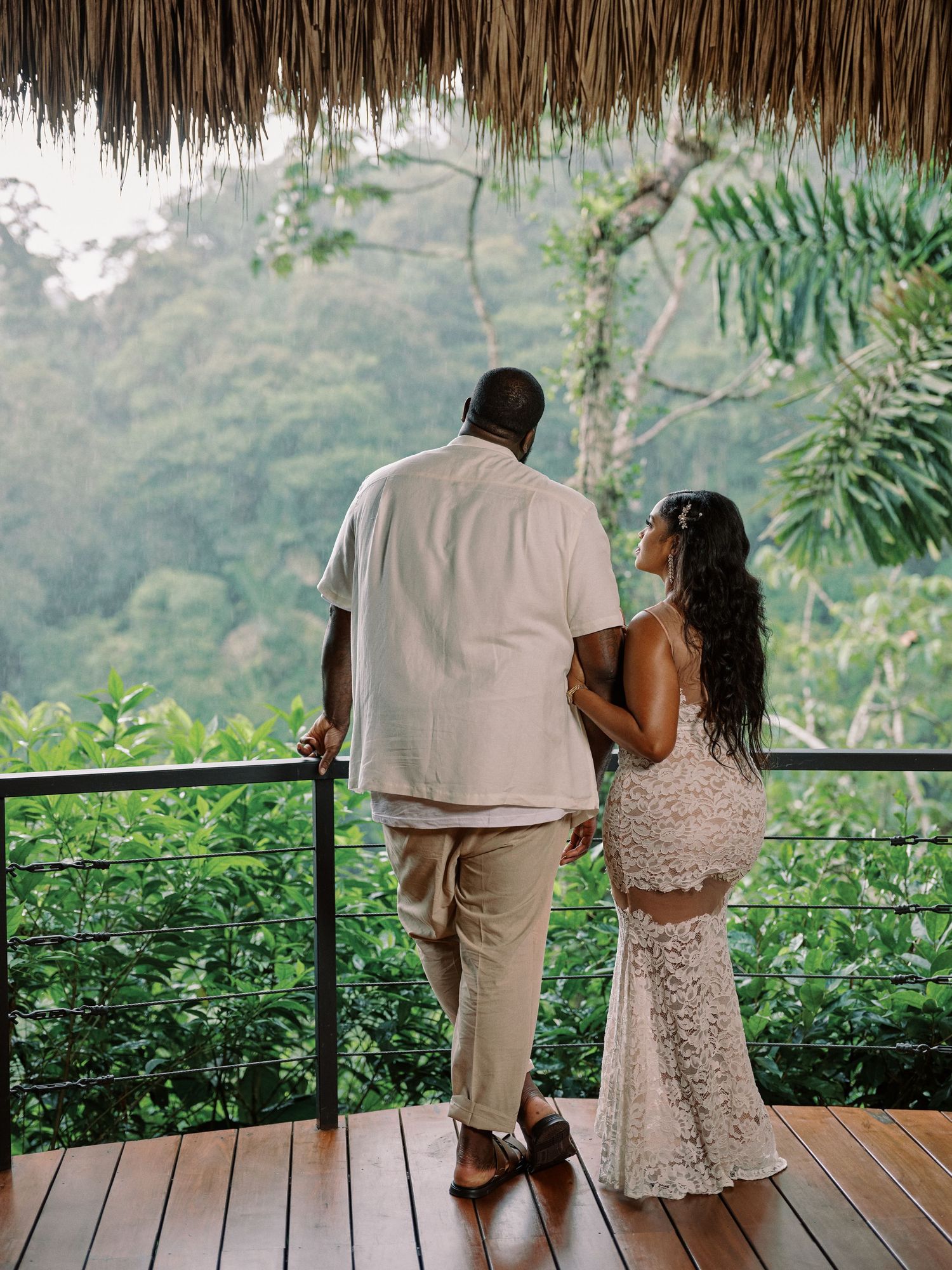 Couple in white attire standing on a tropical wooden deck overlooking lush green jungle scenery.