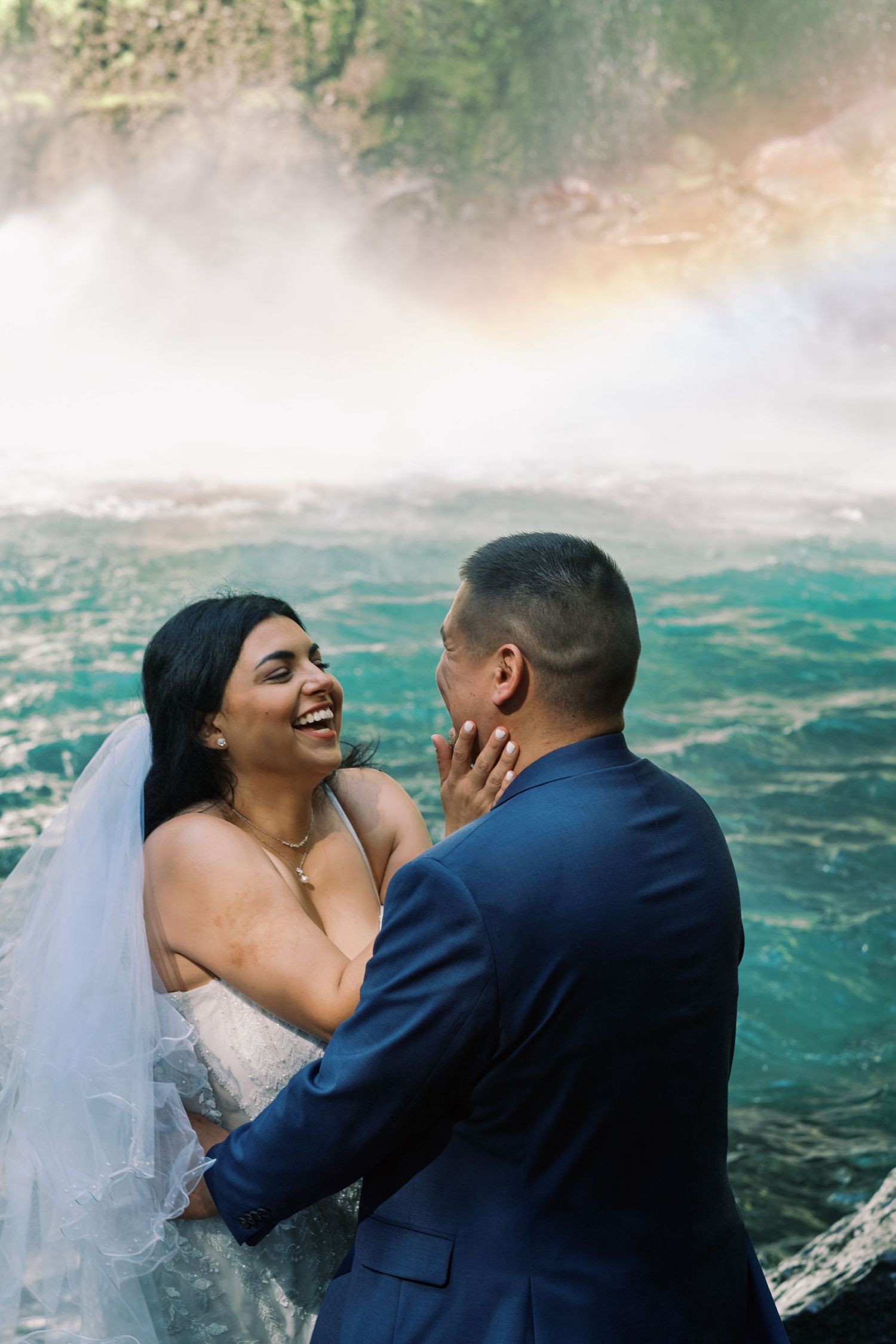 Laughing bride in white gown holds groom's face near a misty waterfall with rainbow backdrop.