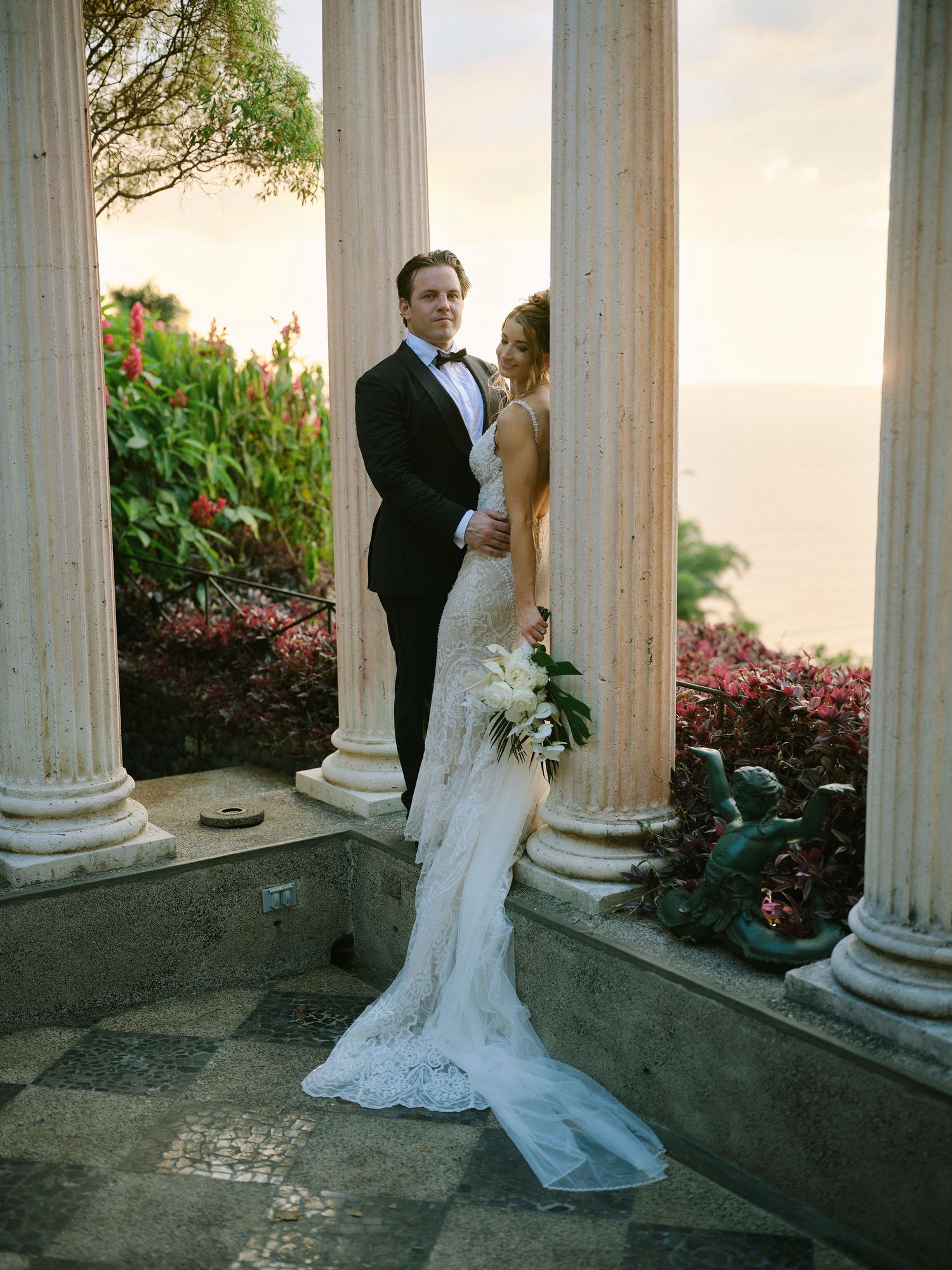 Bride and groom posing between white columns at a tropical cliffside wedding venue at sunset.