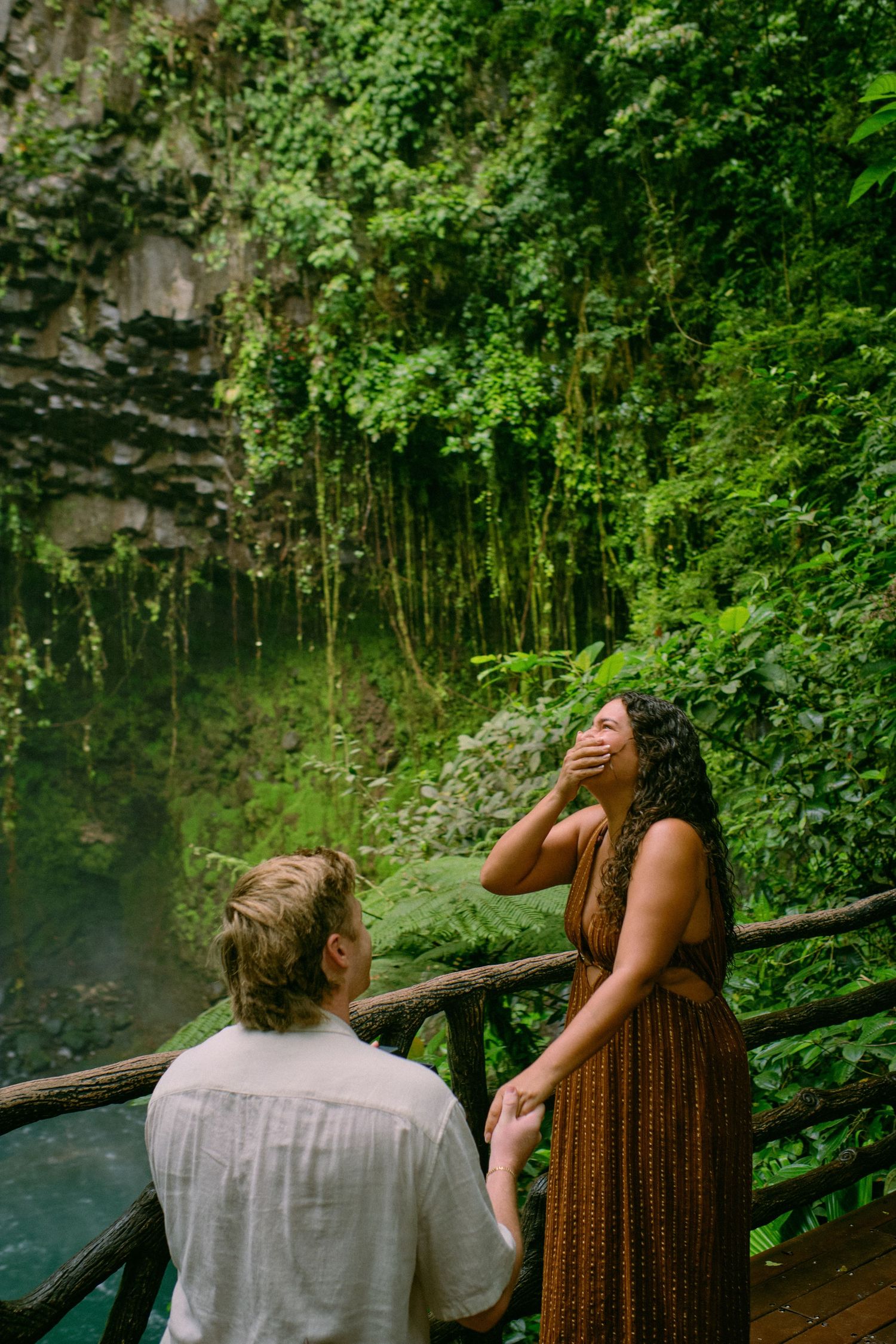 Woman laughing with hand over mouth while man kneels beside her on jungle waterfall viewing platform.
