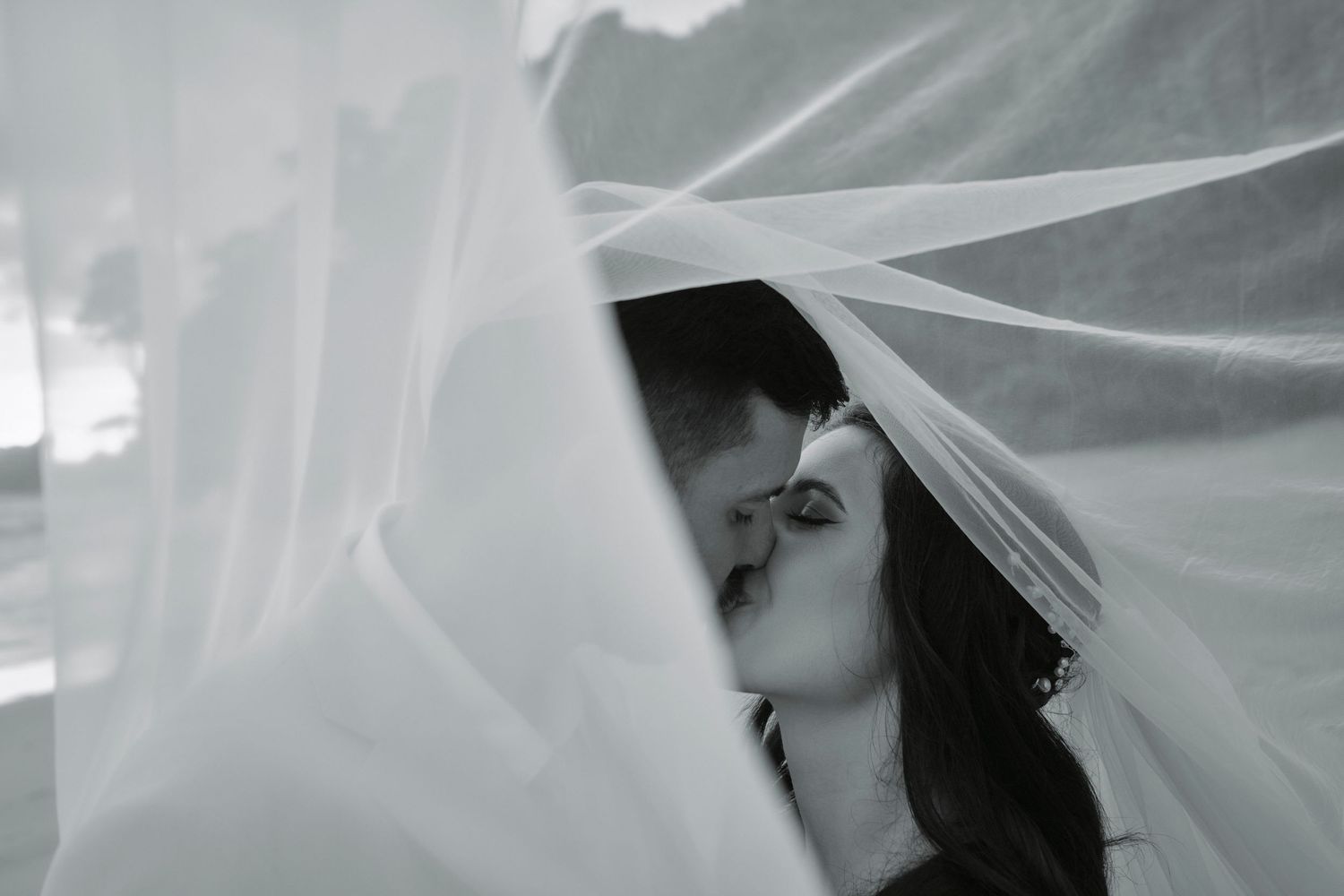 Black and white romantic photo of bride and groom kissing under sheer wedding veil.