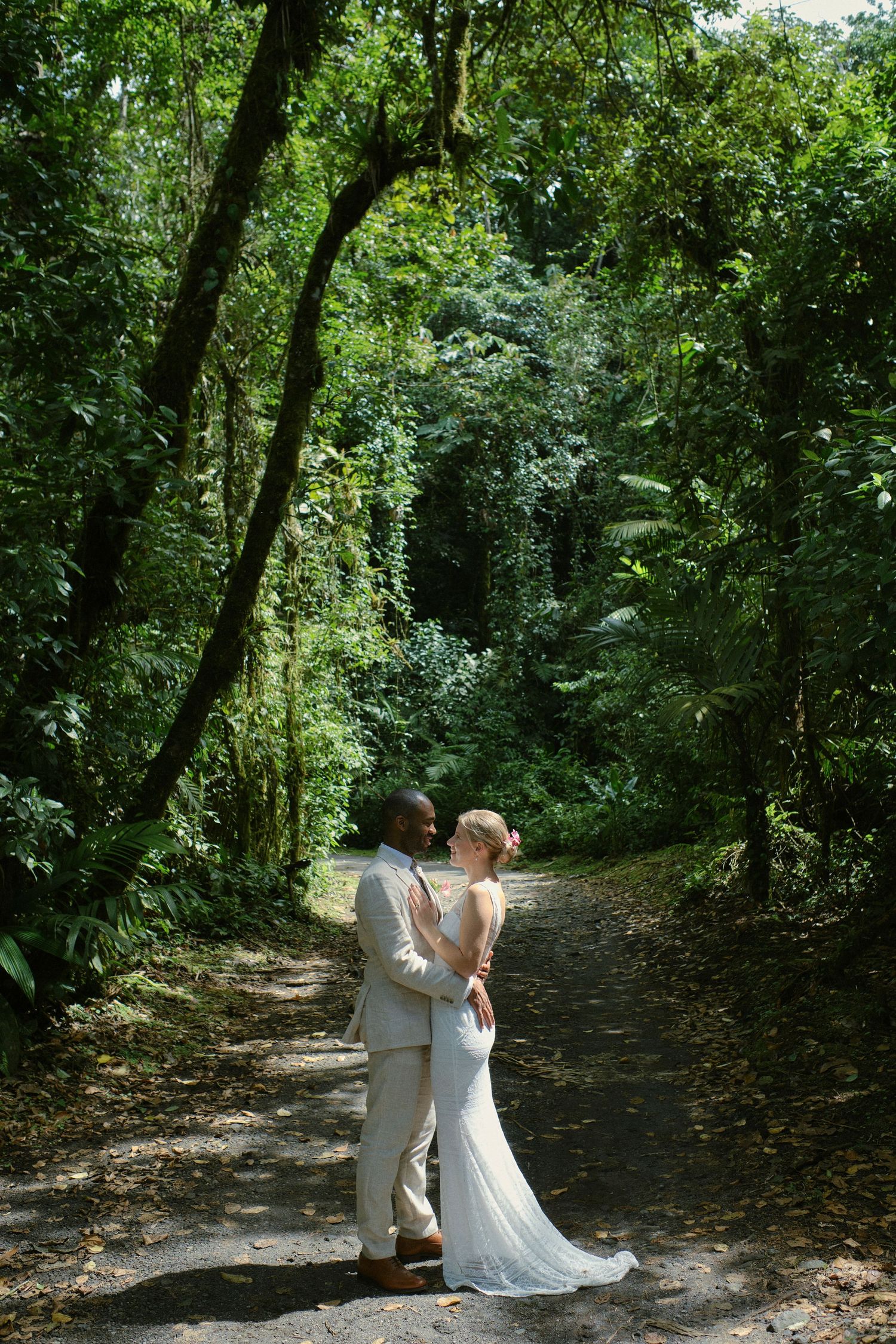Interracial wedding couple embracing on a sunlit path surrounded by lush tropical forest greenery.