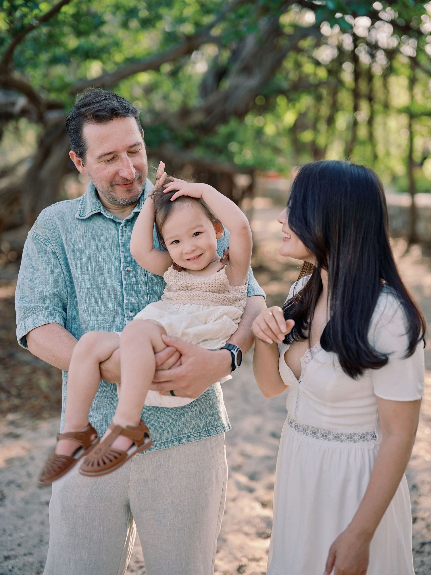 Parents smiling lovingly at their cheerful toddler on a sandy beach path lined with tropical trees.