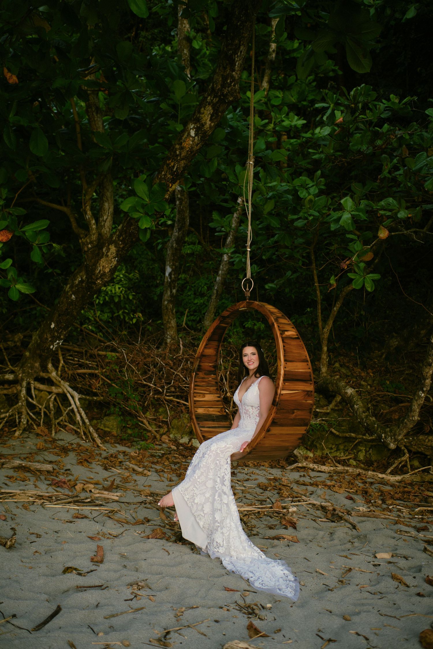 Bride in lace wedding gown sitting gracefully in wooden circular swing on sandy beach.