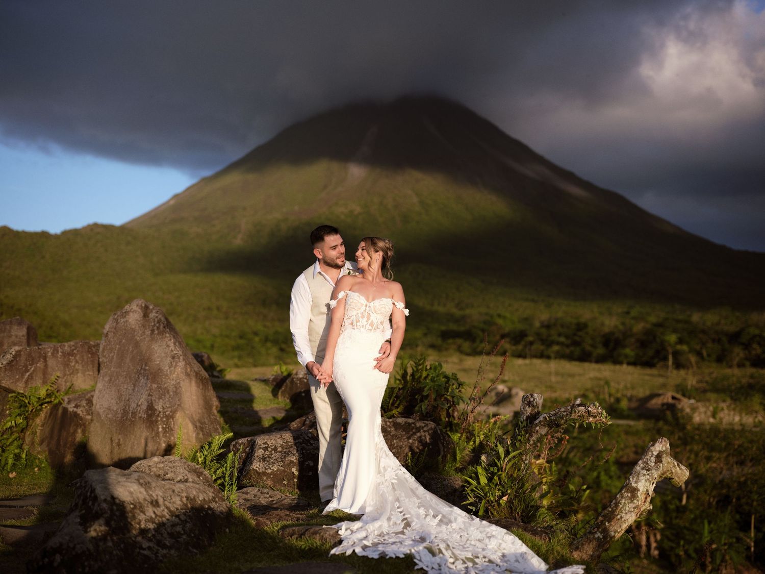 Bride and groom pose dramatically before a volcanic peak with stormy skies and green landscape in Costa Rica.