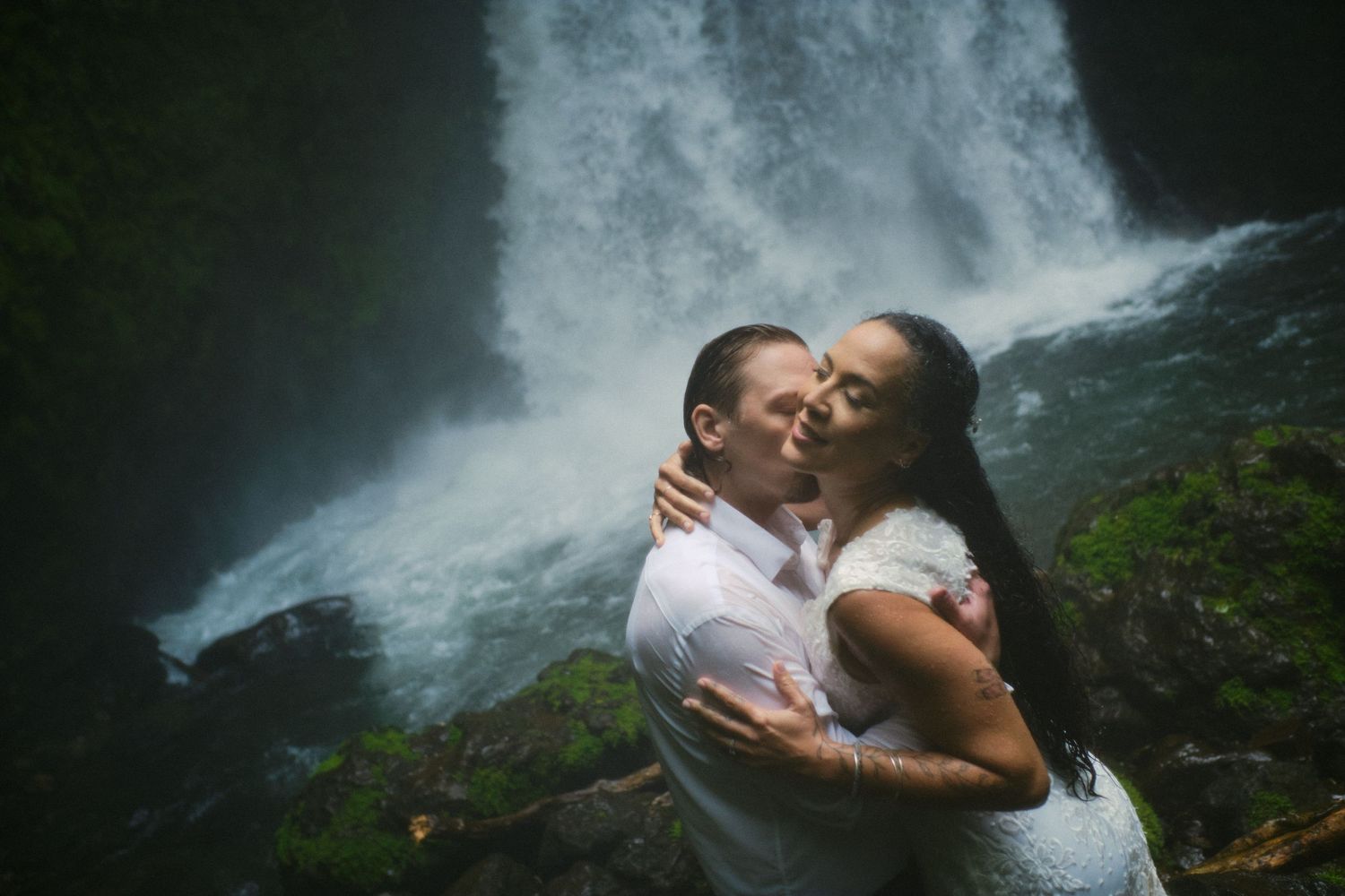 Couple sharing a romantic kiss in front of a stunning waterfall surrounded by lush green foliage.
