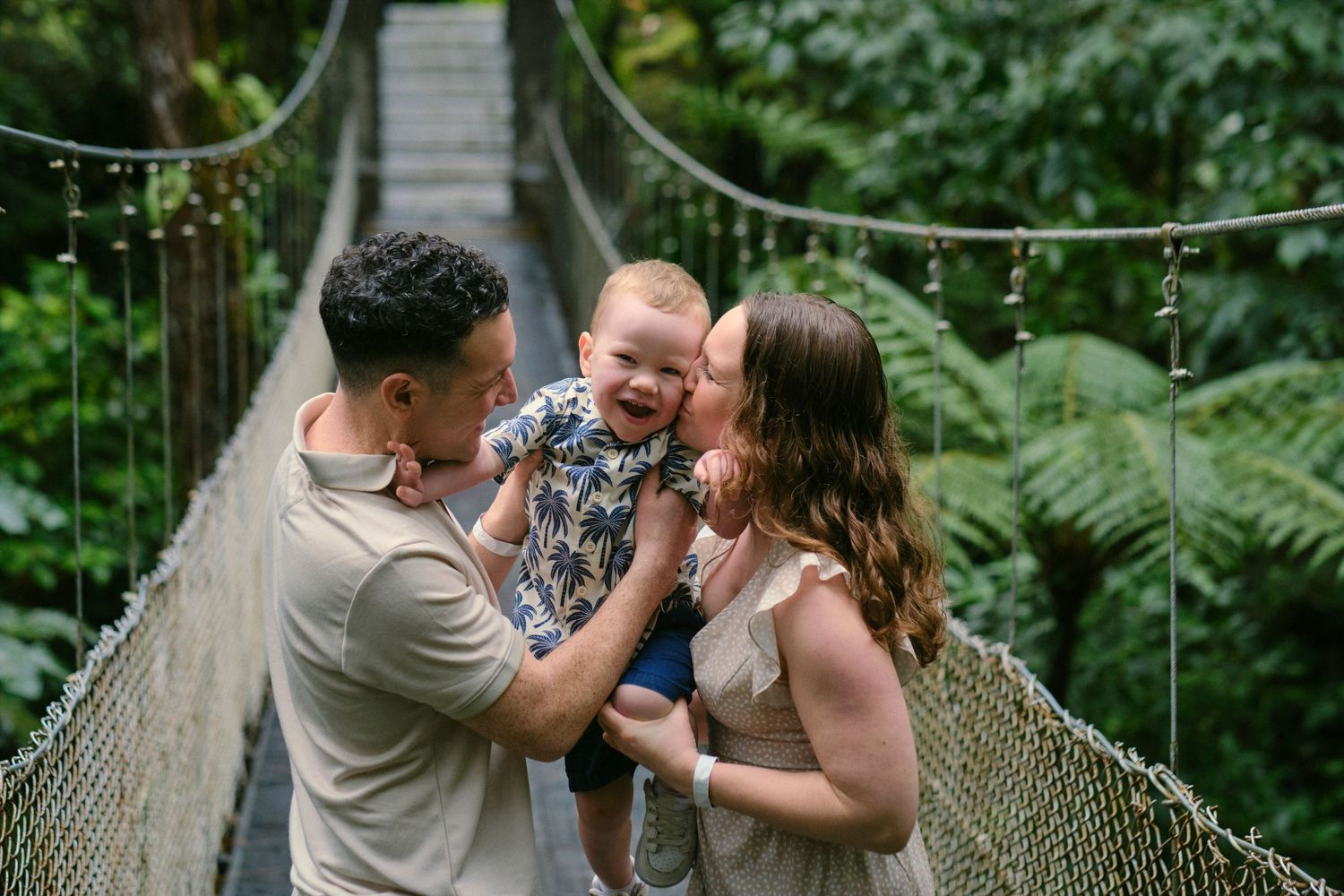 Young couple sharing a joyful moment with their laughing baby on a jungle suspension bridge.