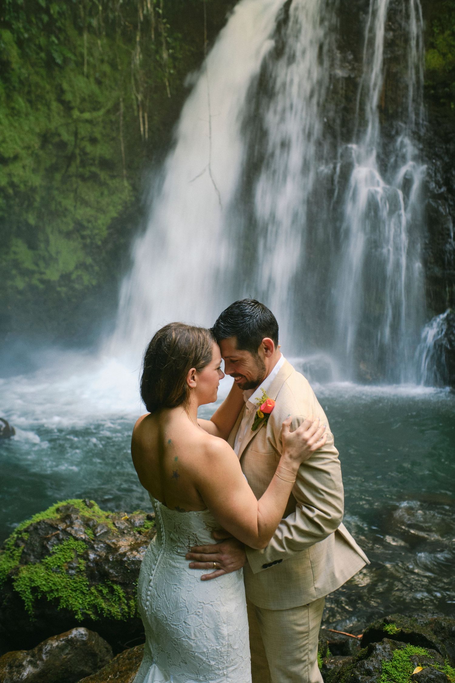 Wedding couple embracing tenderly in front of a cascading waterfall surrounded by lush greenery.