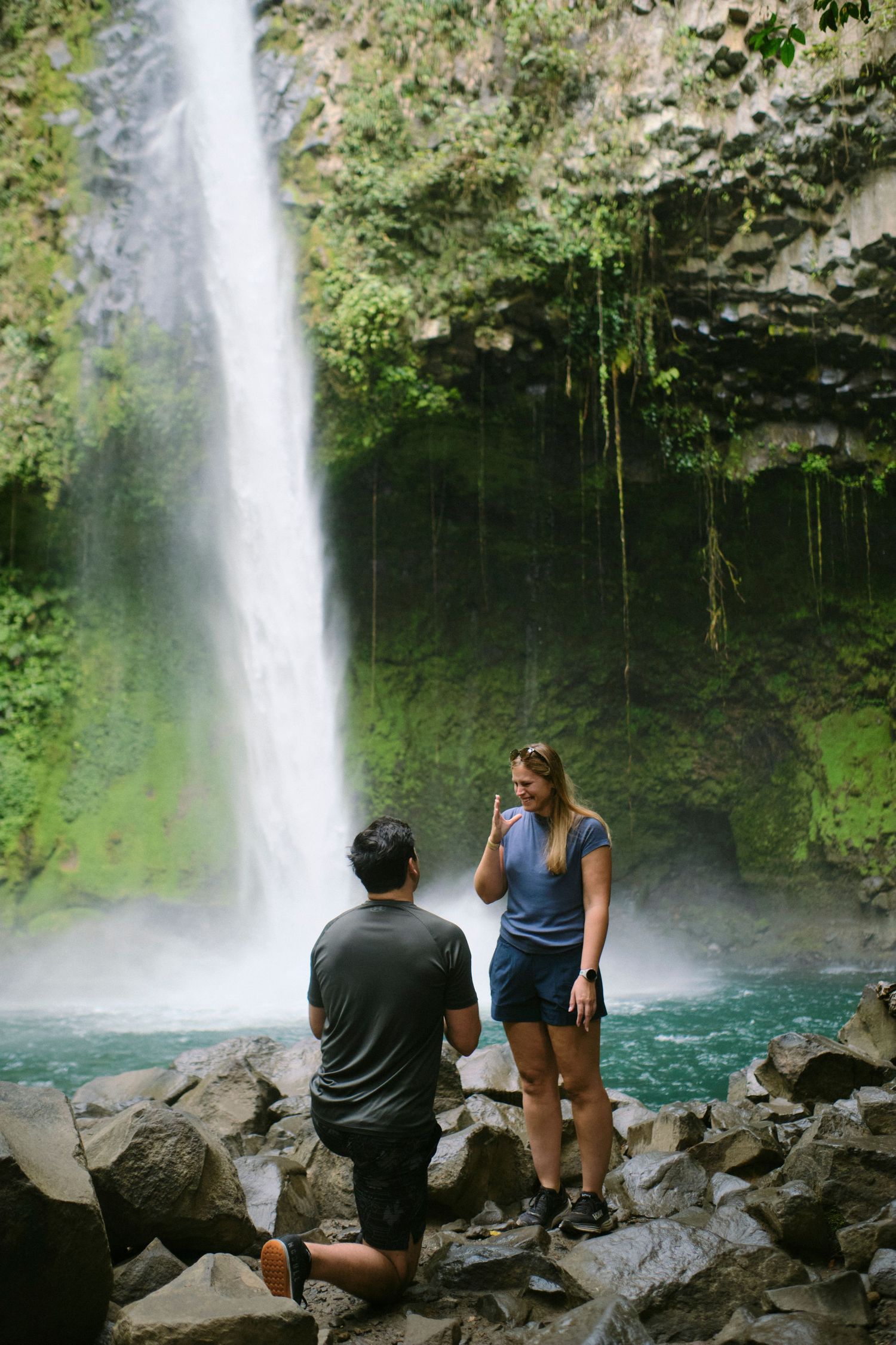 Man proposes on one knee to woman in front of a stunning tall waterfall in lush jungle.
