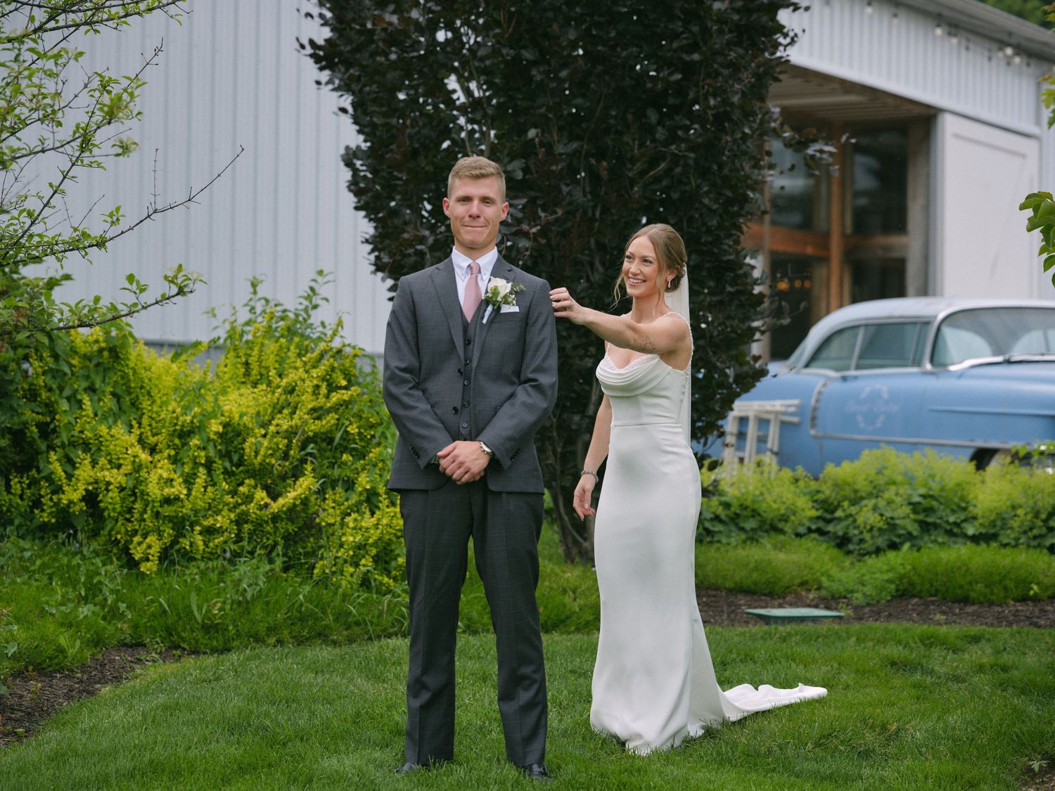 Bride adjusting grooms boutonniere outside a barn venue with a vintage blue car in the background.