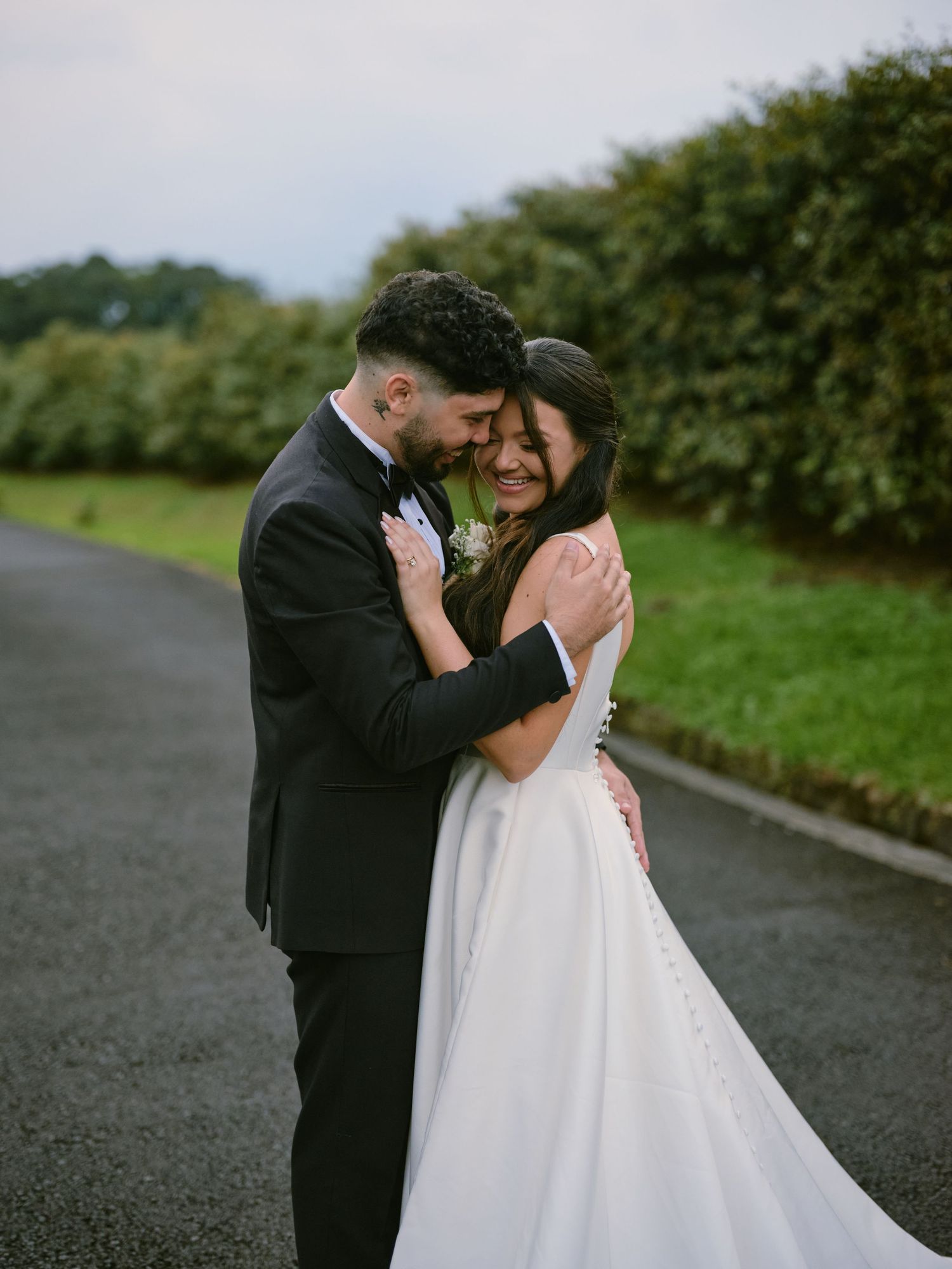 Bride and groom sharing joyful embrace on country road with green hedgerow backdrop.