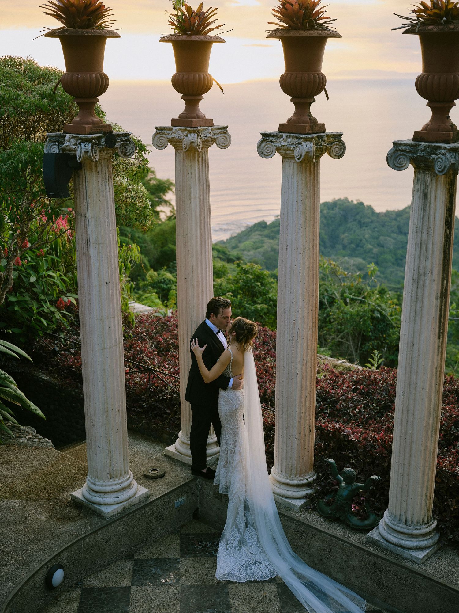 Bride and groom sharing a kiss under ornate columns overlooking a lush tropical coastline at sunset.
